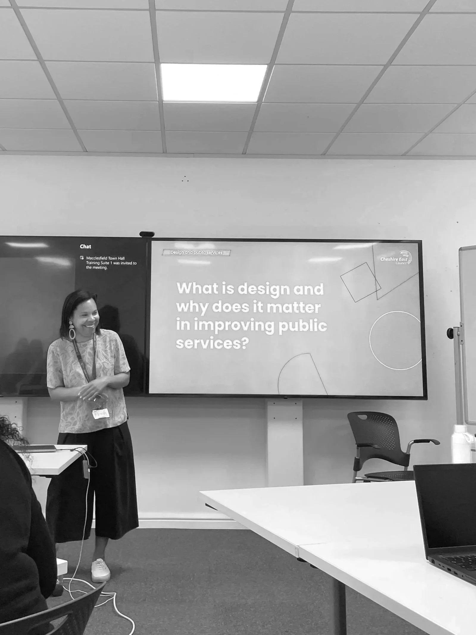 A woman standing in front of a presentation screen with the question, 'What is design and why does it matter in improving public services?' in a classroom setting.
