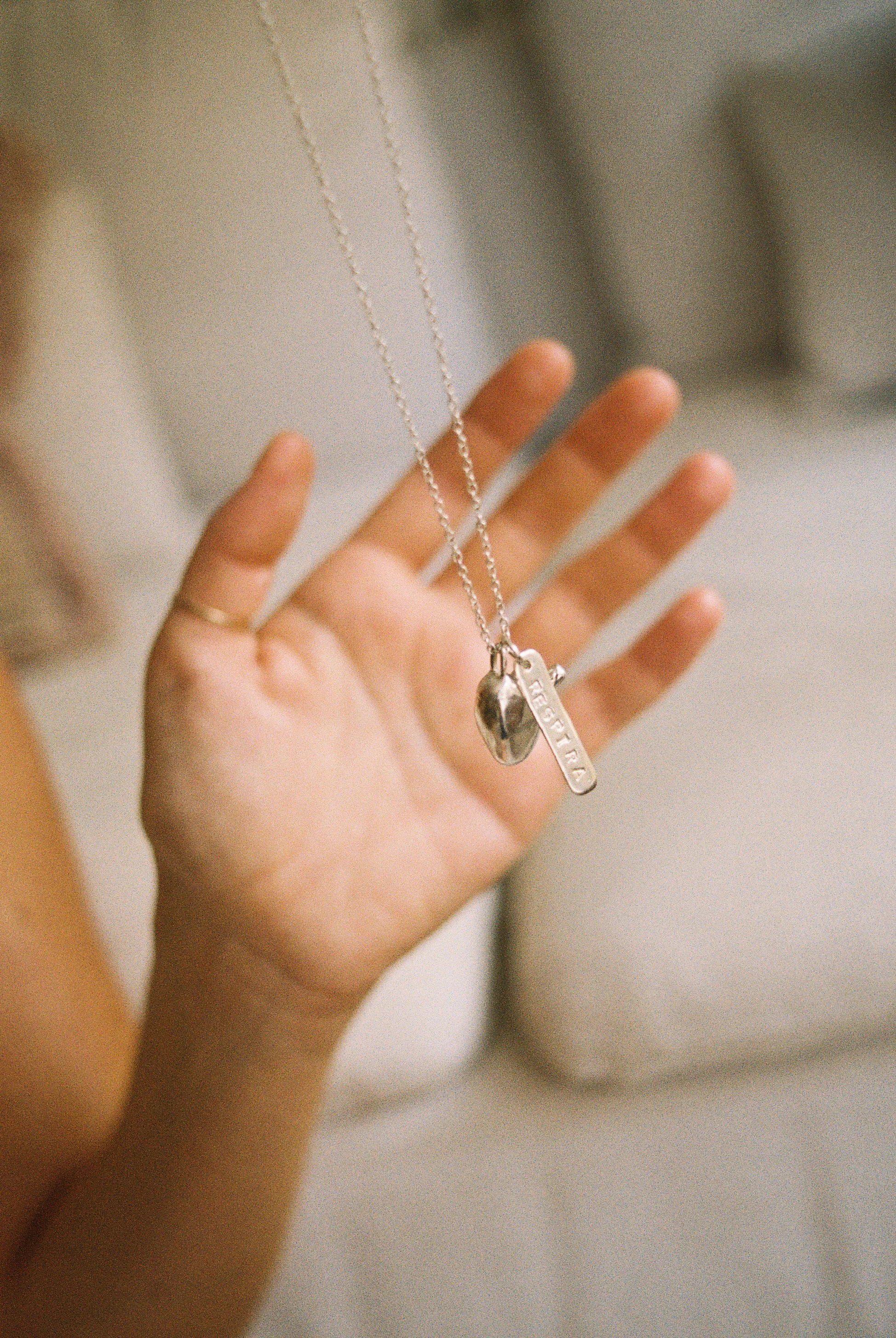 Una mano sostiene tres collares plateados, uno con un dije en forma de corazón, otro con una etiqueta que dice 'IMAGINA' y el último con una pequeña figura. Fondo difuso de tonos neutros.