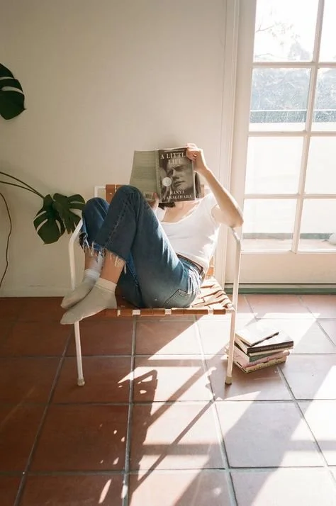 Joven leyendo un libro mientras está sentada en una banca, con luz natural entrando por una ventana grande, en una habitación luminosa con piso de cerámica.