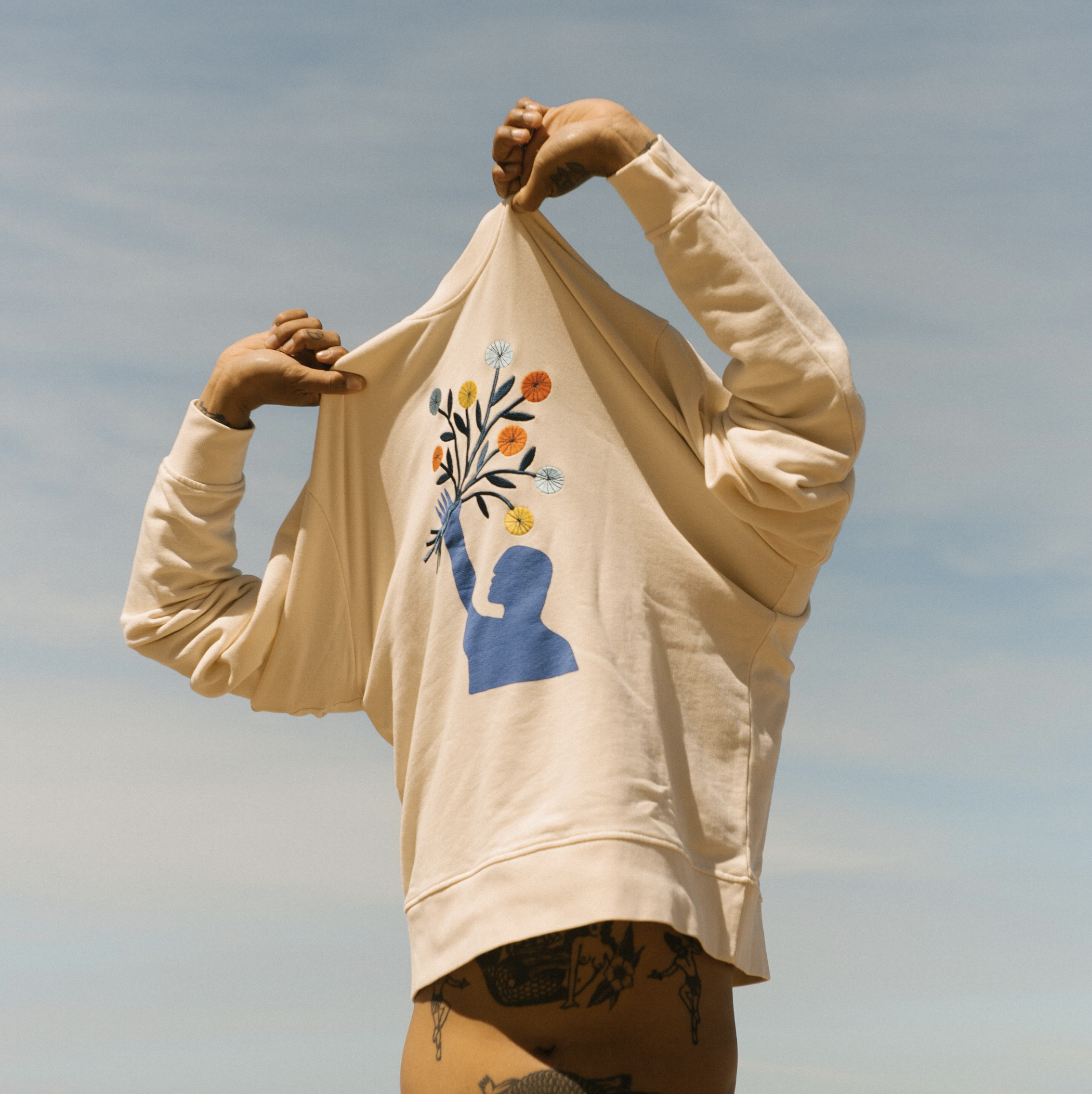 Persona levantando la camiseta para mostrar un diseño con figura de un perfil humano y un árbol con ramas y hojas con flores en diferentes colores.