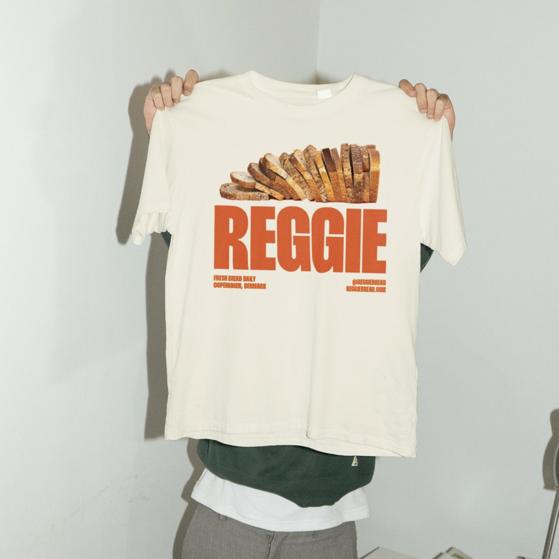 Persona sosteniendo una camiseta blanca con imagen de pan y la palabra 'REGGIE' en letras grandes naranjas.