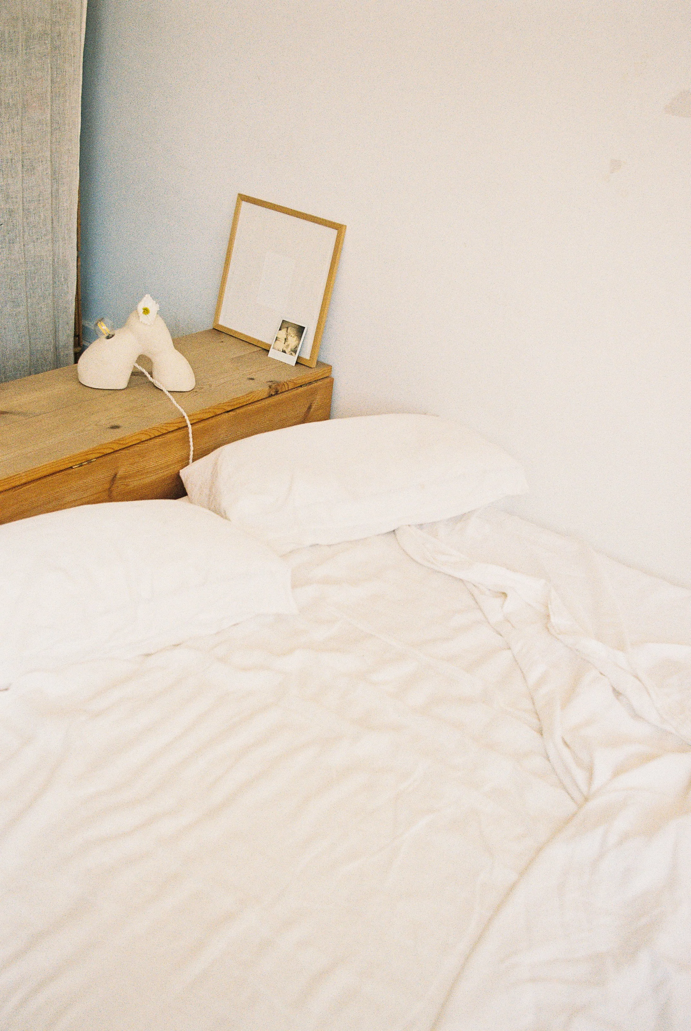 Cama con sábanas blancas, cabecera de madera, lámpara en forma de figura de cerdo, cuadro vacío en la cabecera, y una pequeña fotografía en el mueble al lado.
