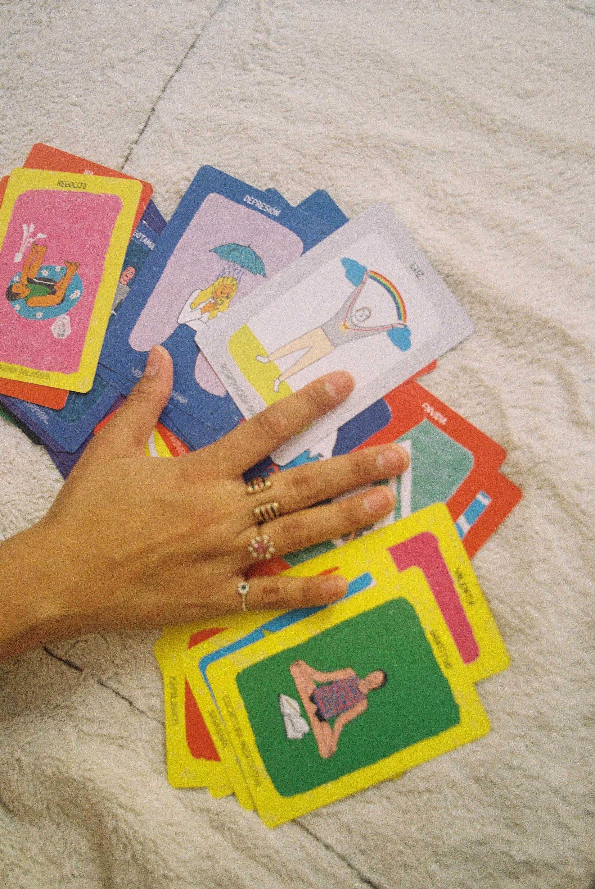 Una mano con varias argollas y anillos sosteniendo un conjunto de cartas de juegos de cartas en diferentes colores y con ilustraciones y palabras en español, sobre una superficie de tela clara.