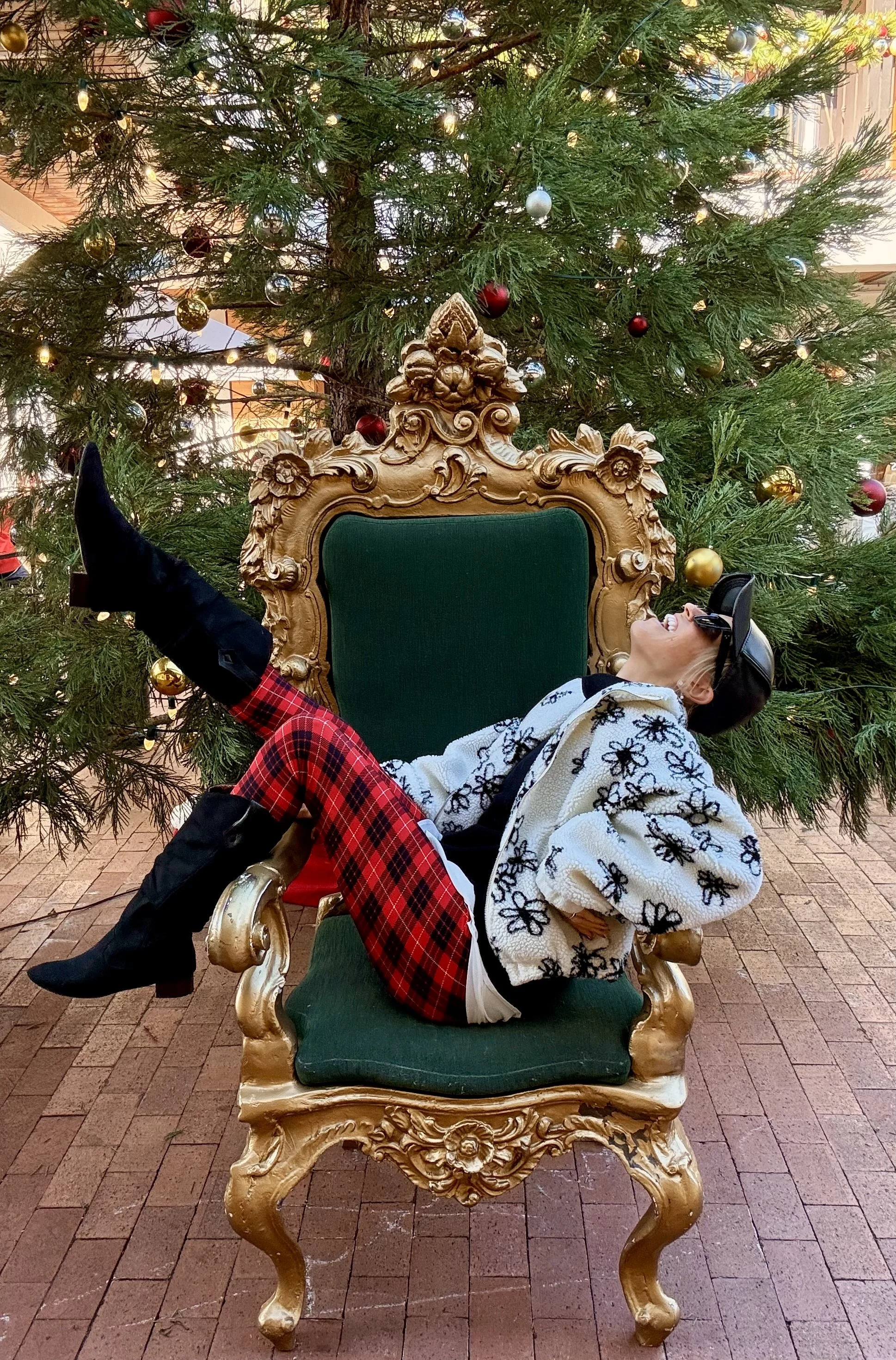 Person lying on a green and gold ornate throne in front of a decorated Christmas tree, smiling and wearing sunglasses, a hat, and colorful clothing.