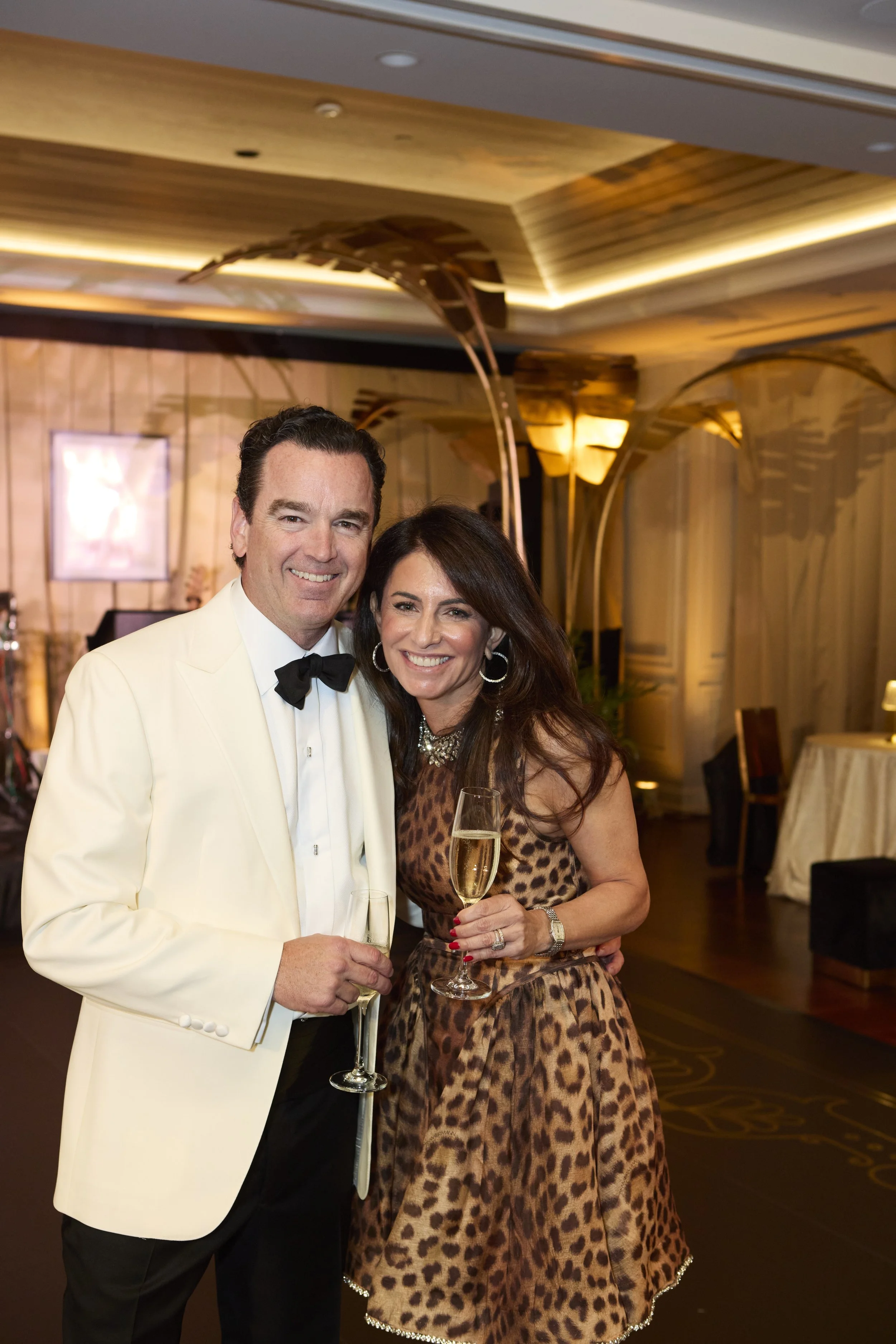 A man in a white tuxedo with a black bow tie and a woman in a sleeveless leopard print dress smiling and holding glasses of champagne, standing in an elegant, warmly lit event space.