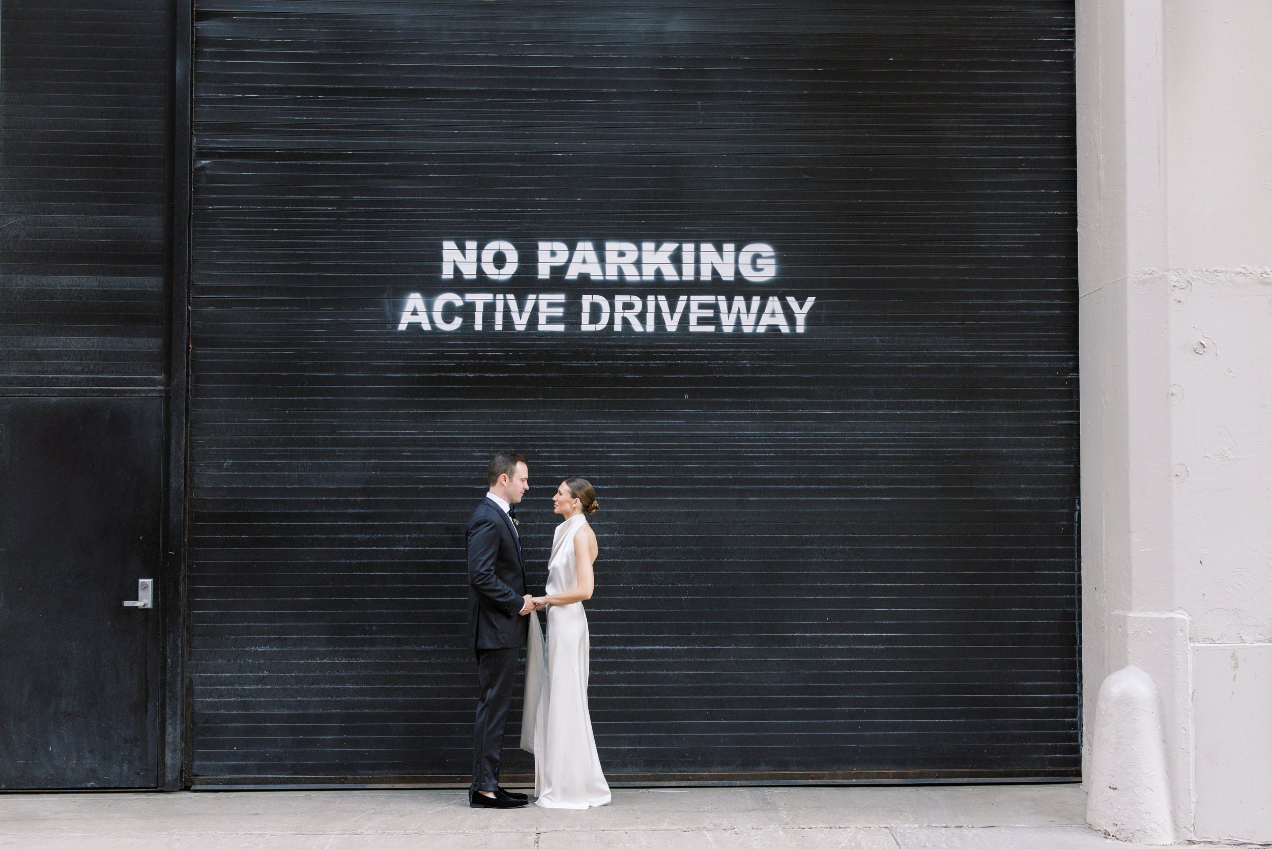 A couple in formal attire, a man in a suit and a woman in a white dress, stand facing each other holding hands in front of a large black garage door with a sign that reads 'NO PARKING ACTIVE DRIVEWAY'.