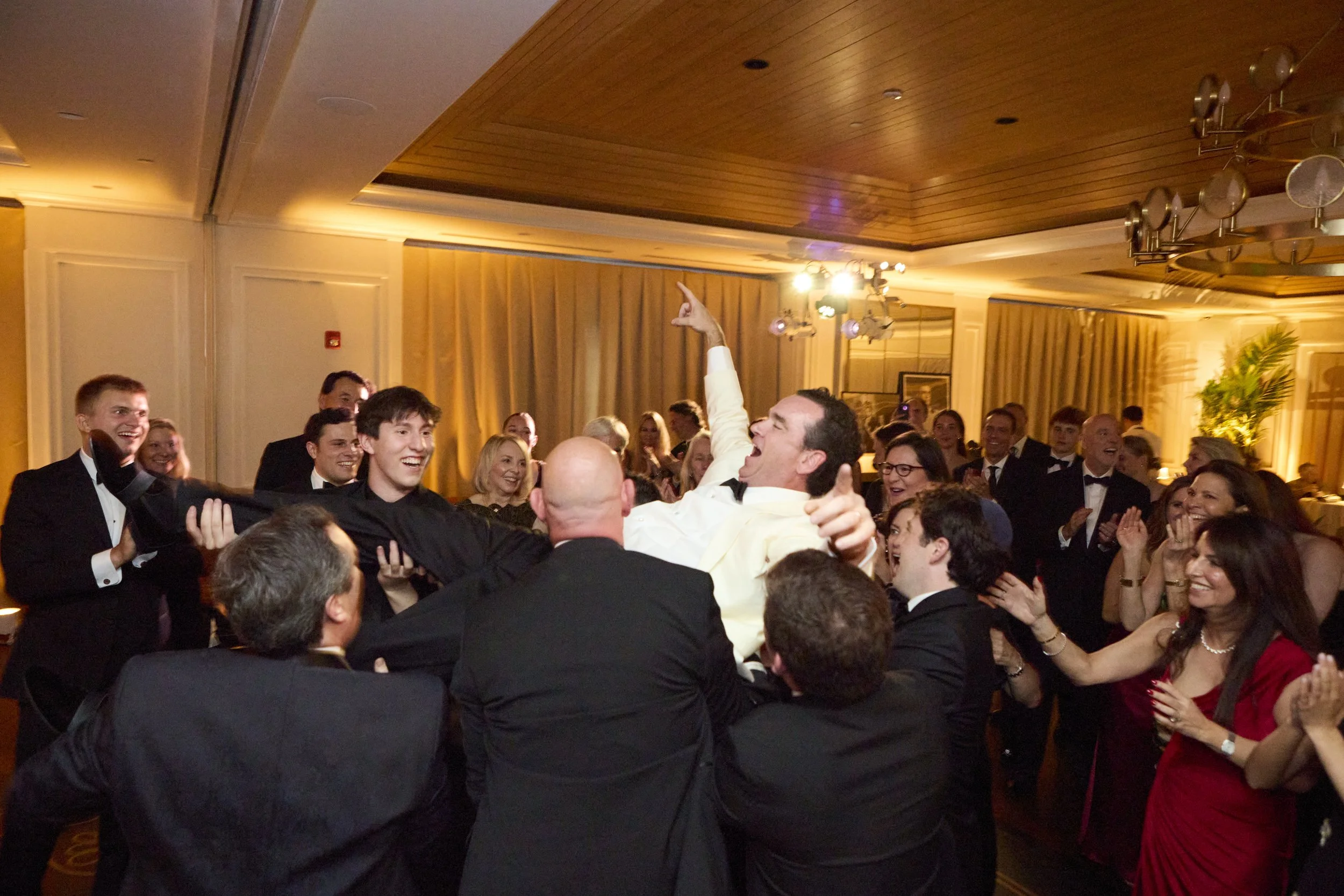 A lively celebration with people in formal attire dancing and cheering indoors, with a man lifted and surrounded by friends.
