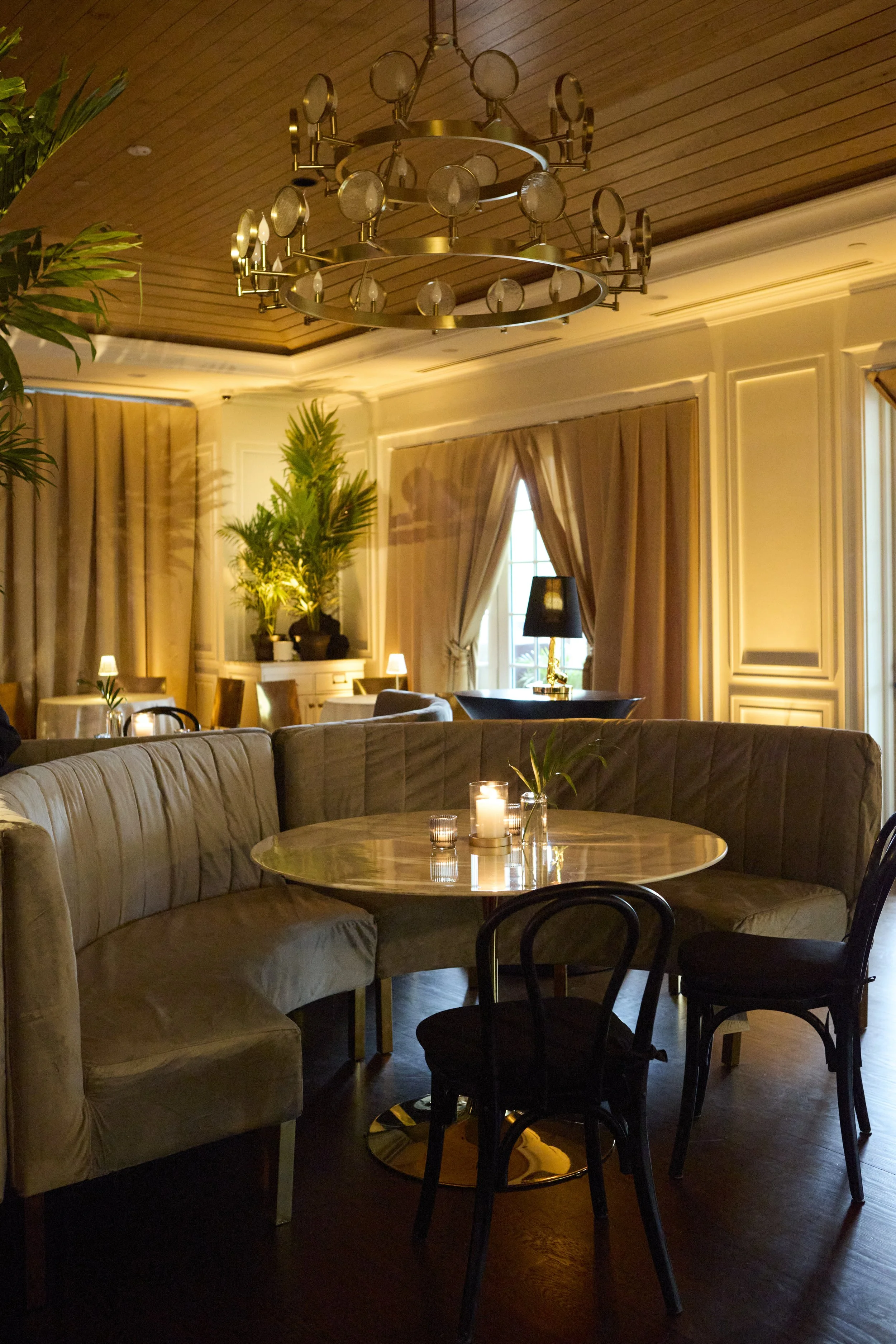 Elegant dining area with a round marble table, candle, and vases, surrounded by velvet and wooden chairs, with draped curtains, lush potted plants, and a modern chandelier hanging from a wooden ceiling.