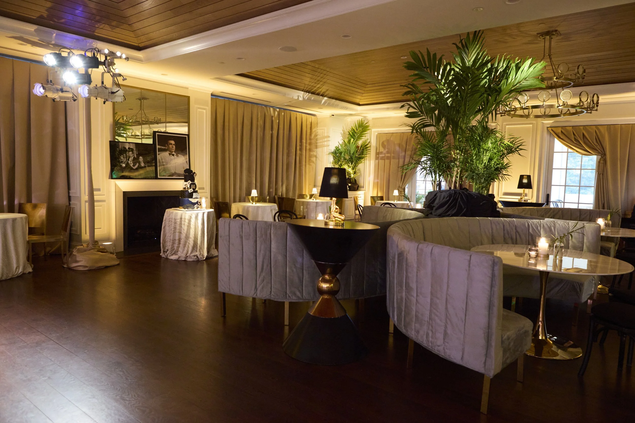 An elegantly decorated lounge with seating, plants, candles, table lamps, and framed photographs, illuminated by warm lighting and large windows with curtains.