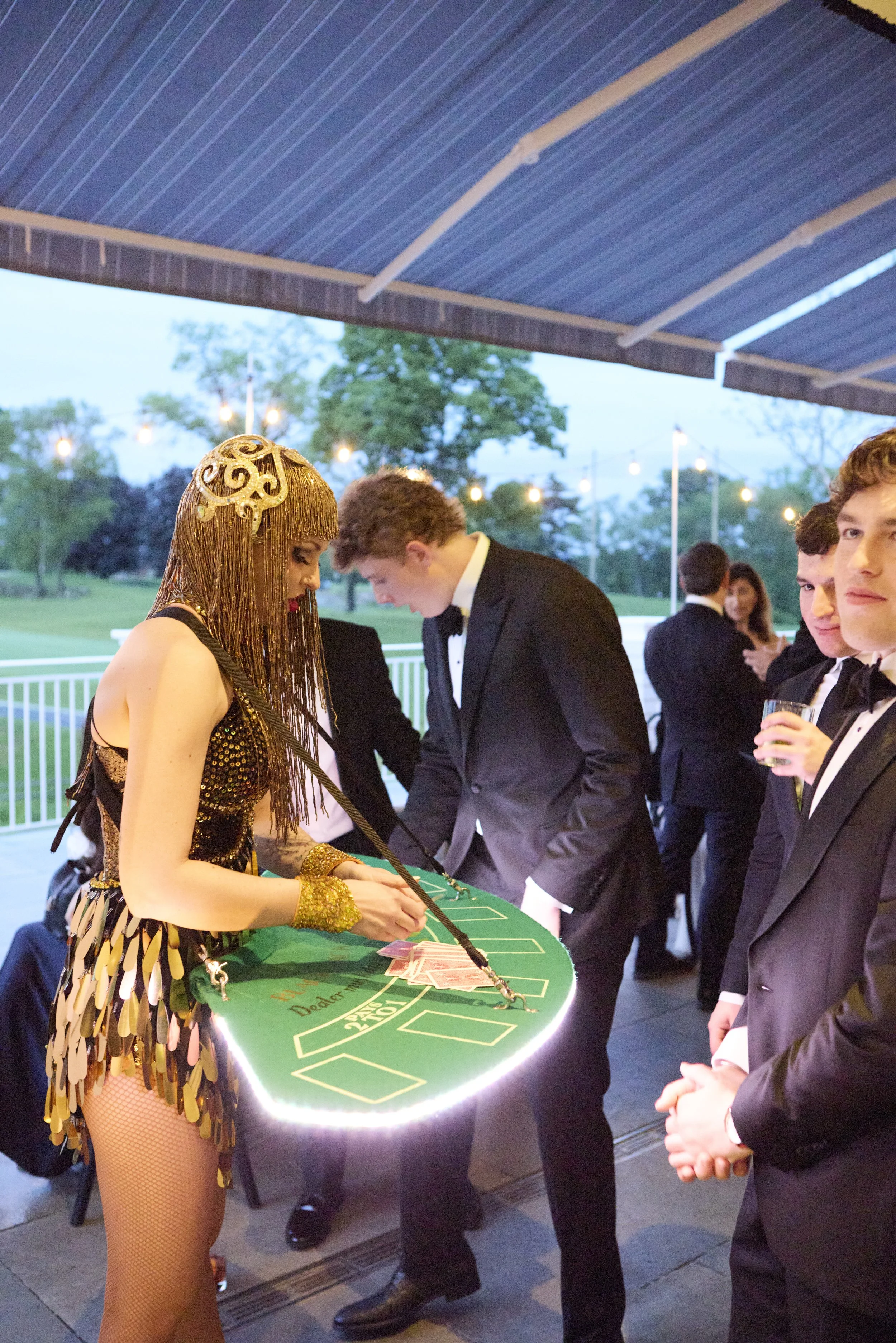 Young woman dressed in a gold and black costume with gold accessories, including fringe and gloves, playing roulette at an outdoor casino party with party guests in tuxedos watching.