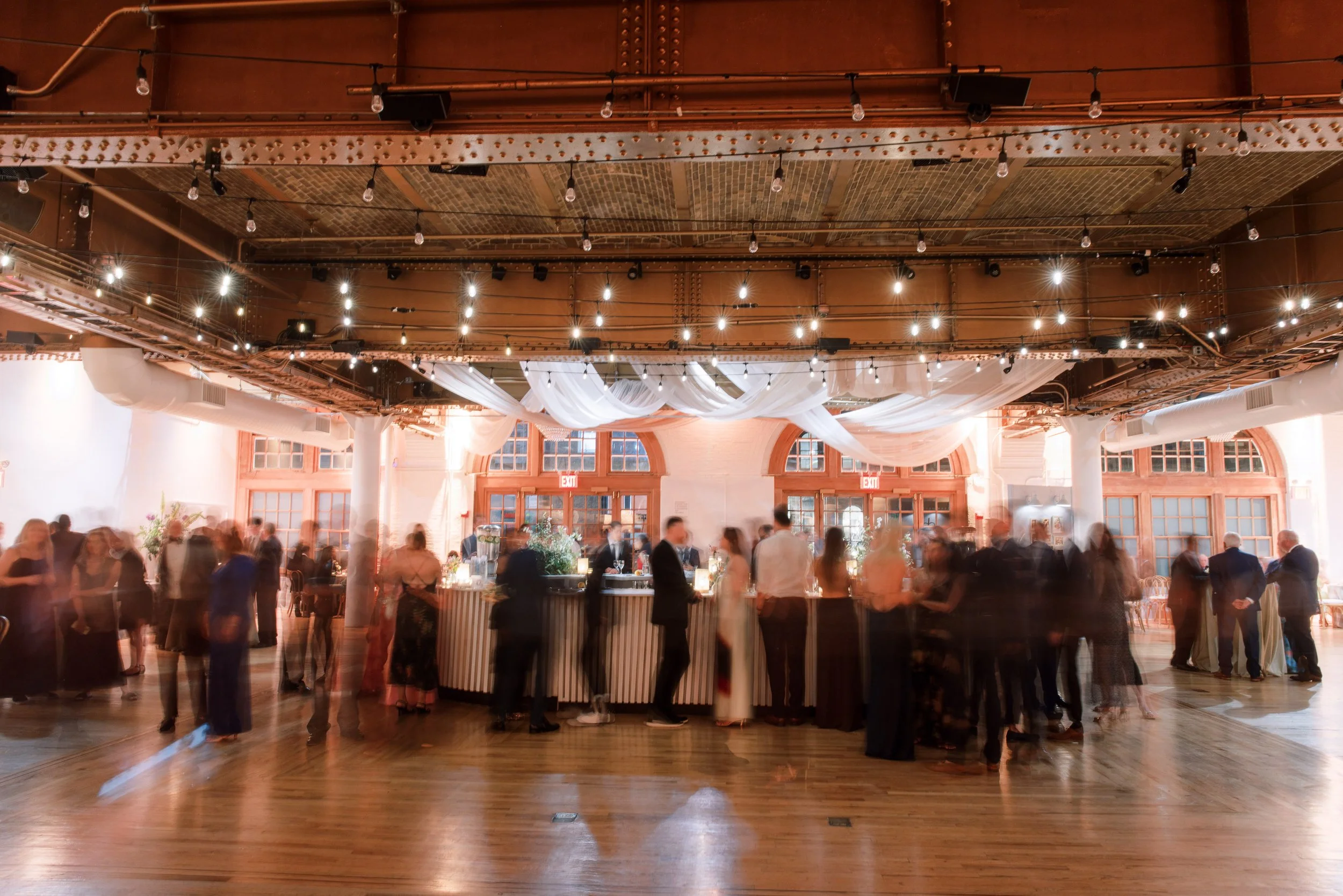 An indoor event space with string lights, draped white fabric, and large windows. People are gathered around a bar and in small groups, with the overall scene showing a social gathering or party.