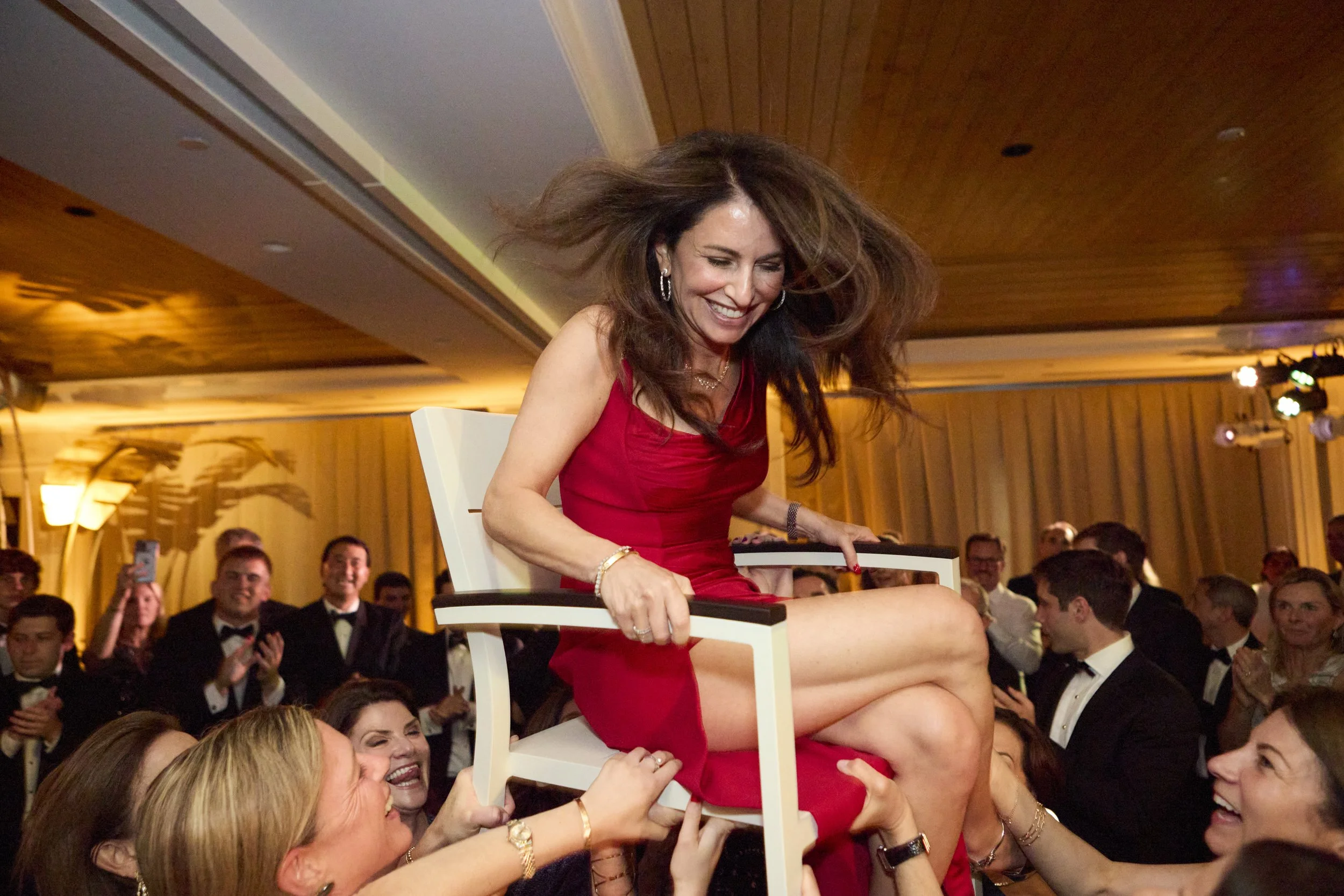 Woman in a red dress sitting on a chair being lifted in the air by a group of people at a celebration or gathering.