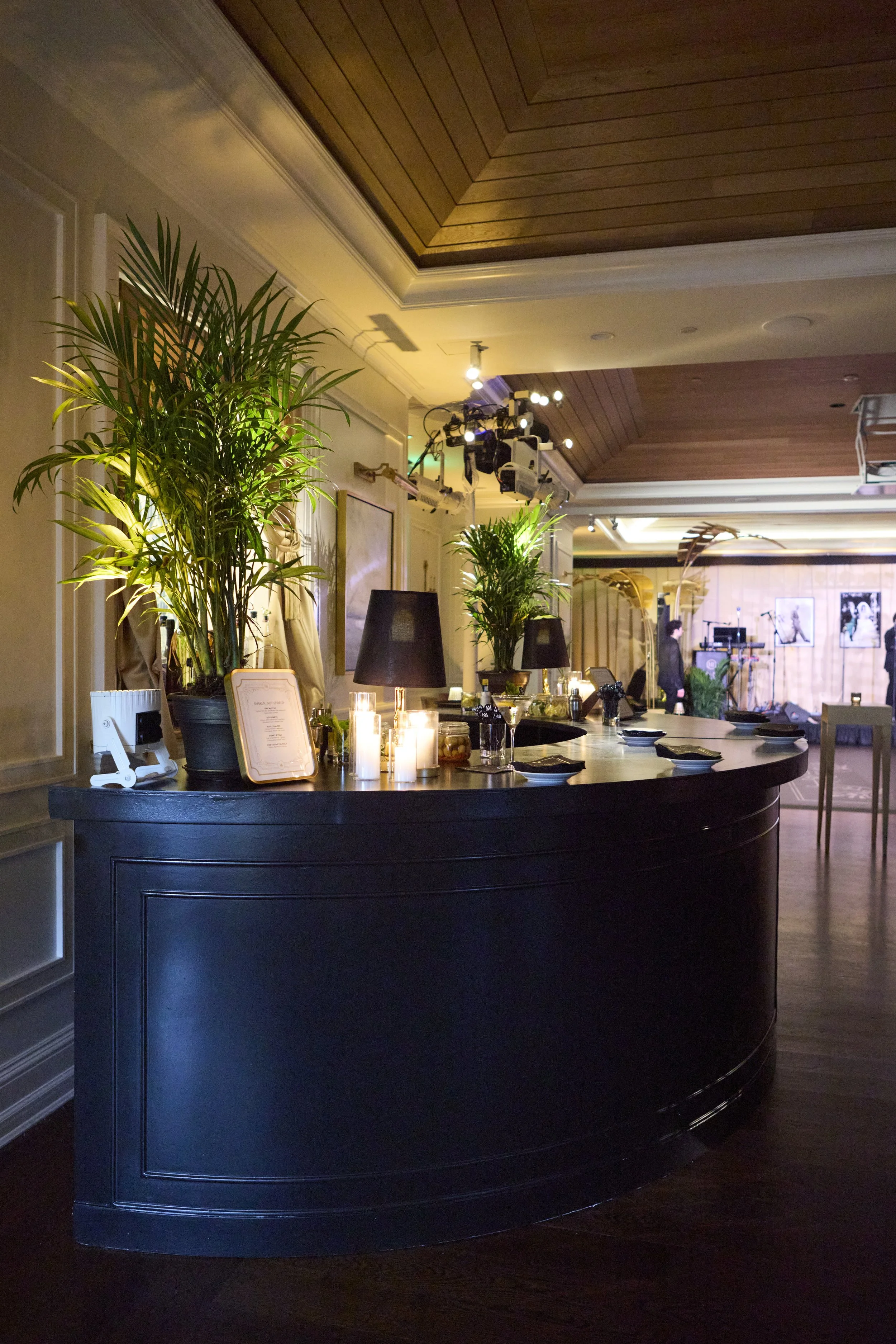 A reception area with a curved black counter decorated with candles, potted plants, and a framed menu. The background has warm wooden panels, framed artwork, and people near a stage with musical instruments.
