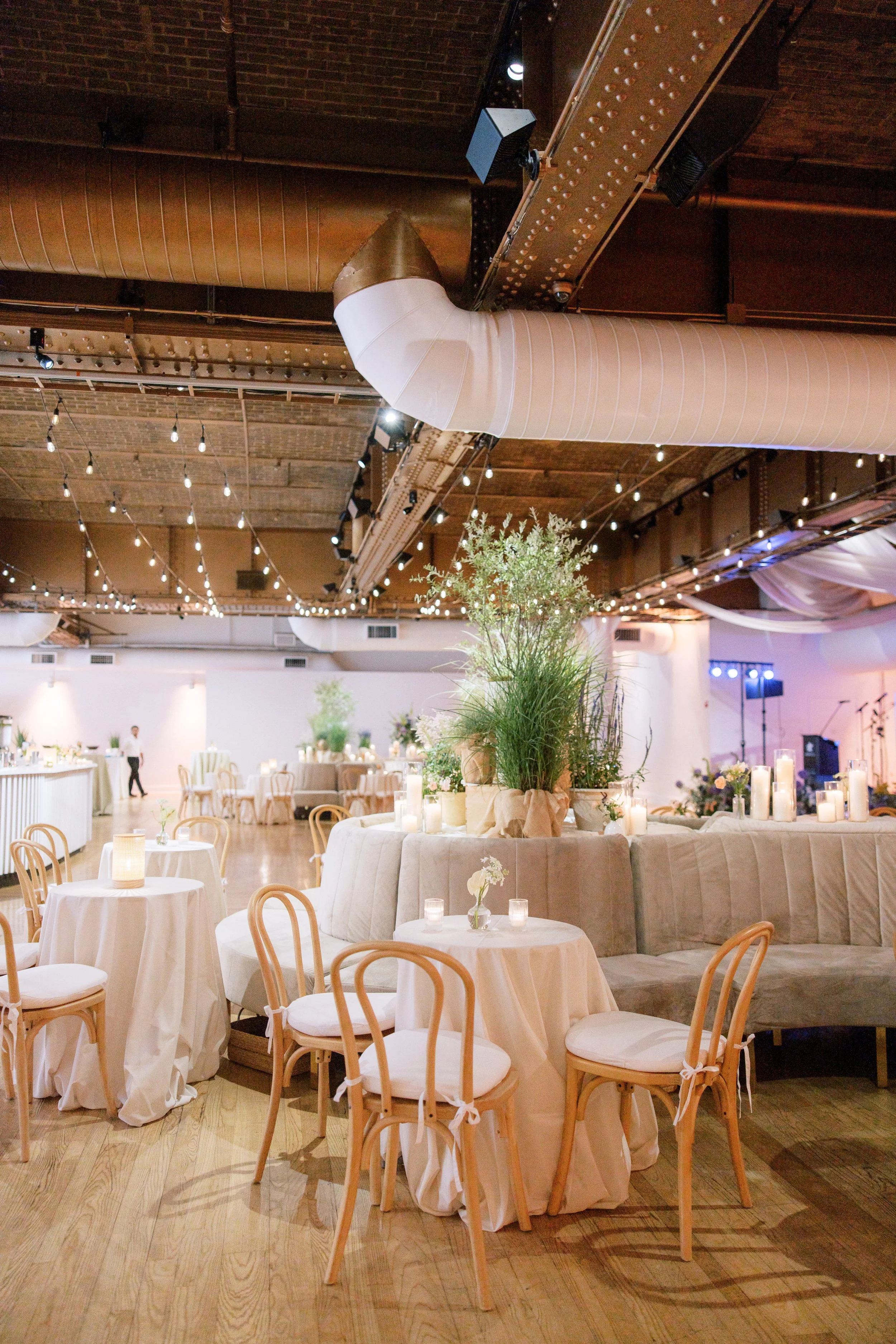 Elegant wedding reception hall decorated with candles, flowers, and white tablecloths, featuring string lights and industrial-style ceiling ductwork.