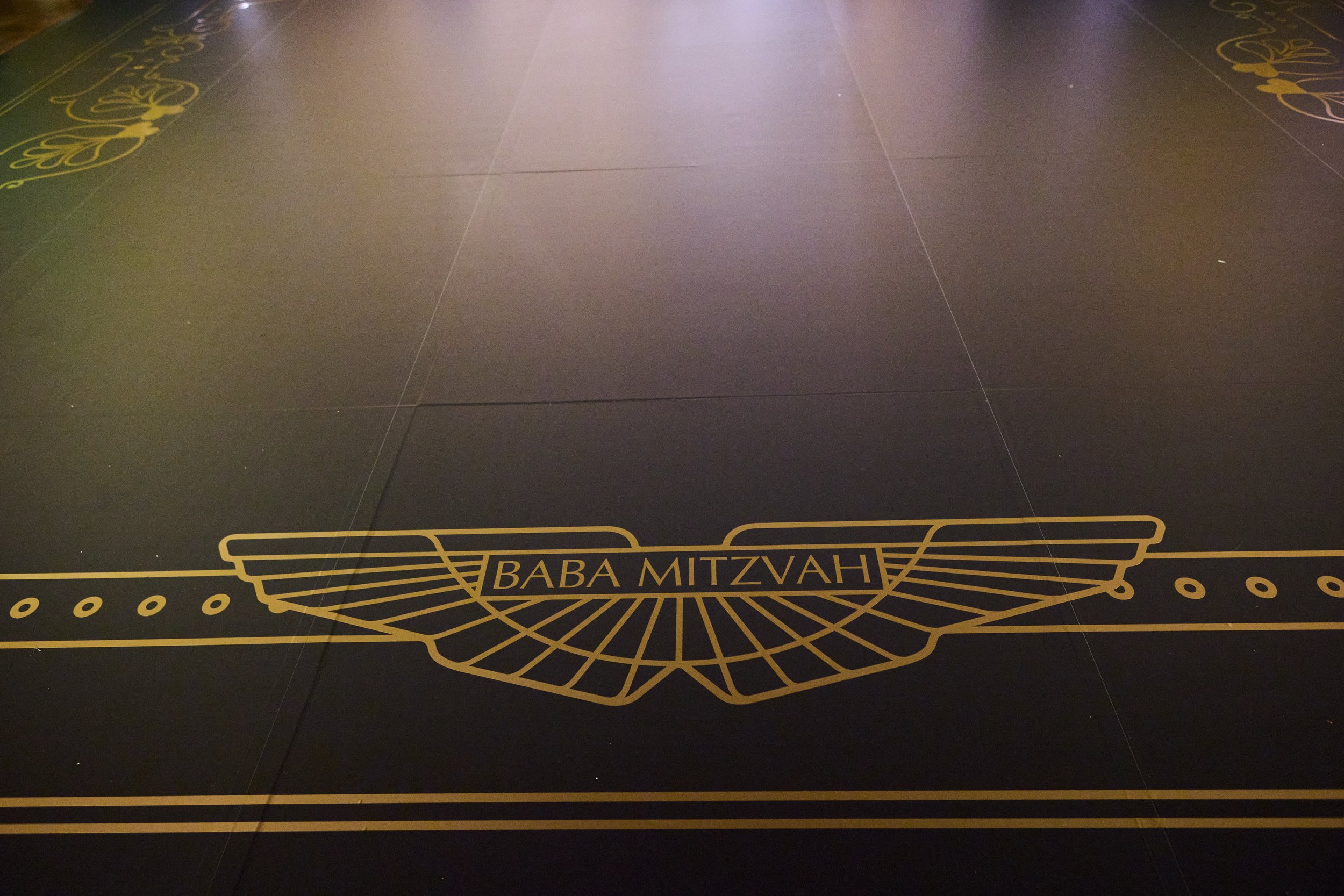 Decorative black floor with gold lines and the words 'BABA MITZVAH' in the center surrounded by a winged design.