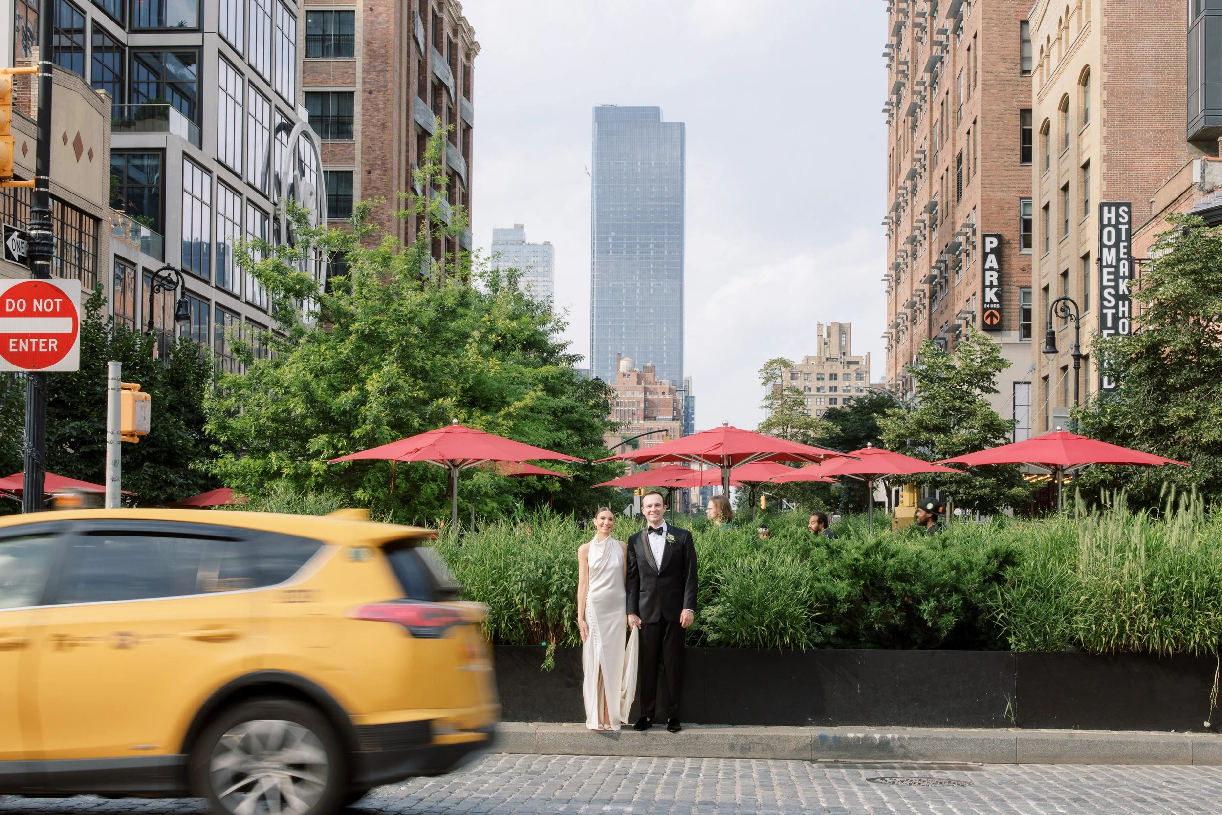 A couple dressed in formal attire, standing on the sidewalk near a lush green garden with pink umbrellas in an urban cityscape. A yellow taxi passes by on the street.