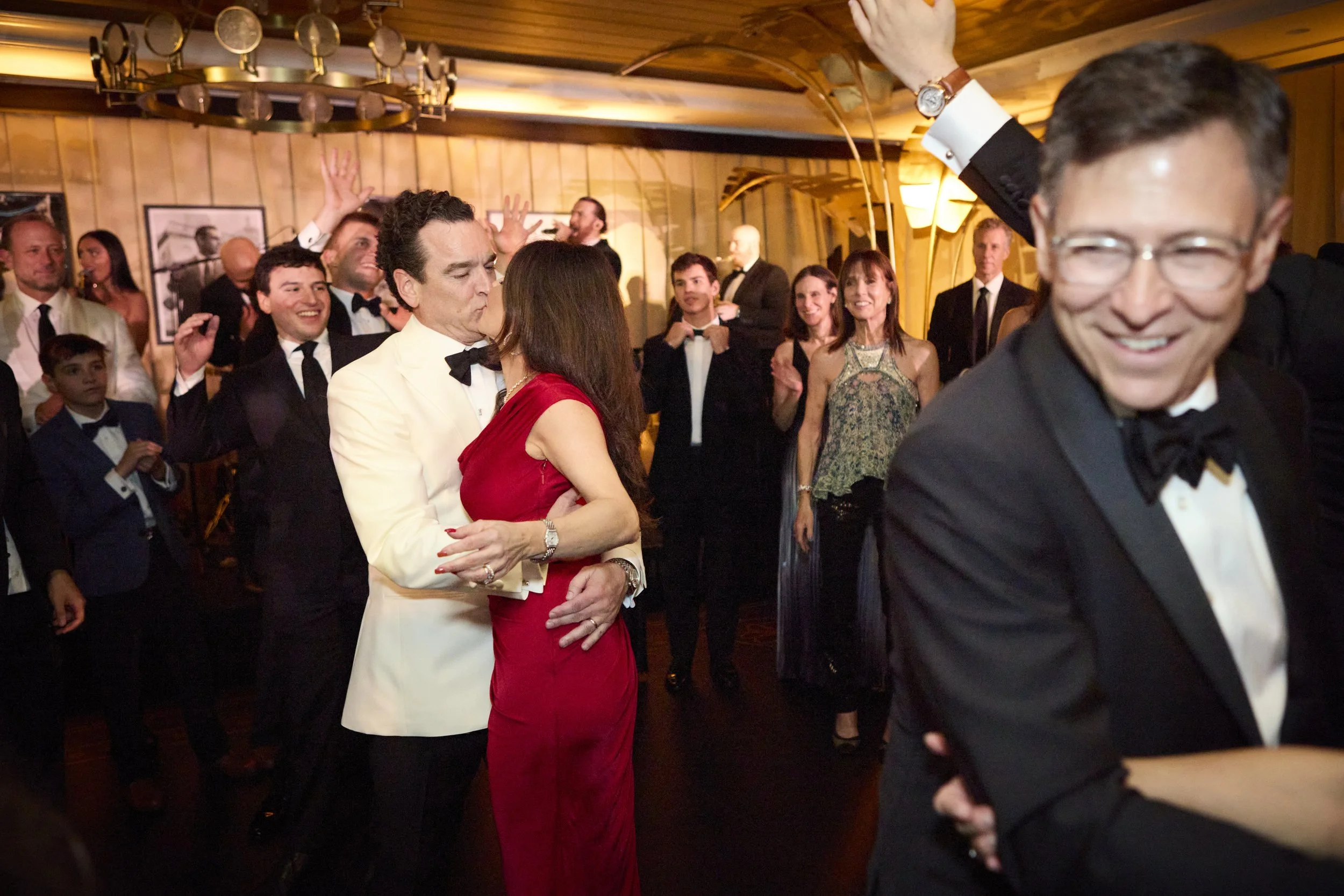 People dancing and enjoying themselves at a formal event or party, with a couple sharing a kiss in the foreground.
