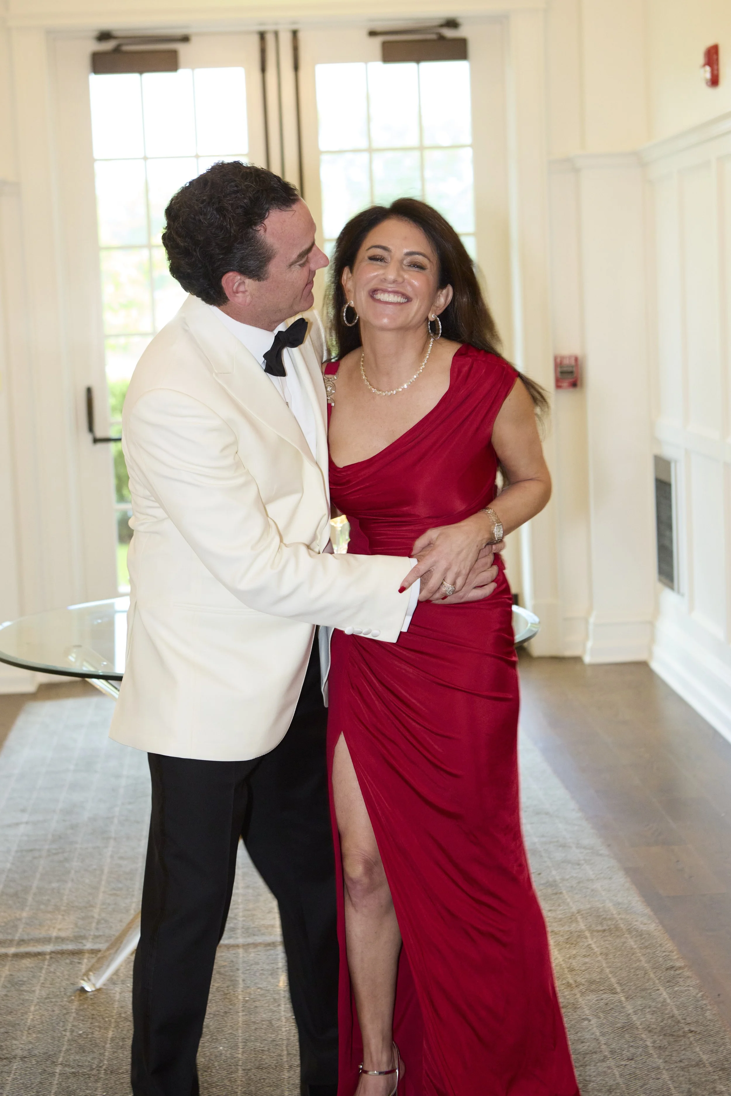 A man in a white tuxedo with a black bowtie and a woman in a red evening gown with a high slit, happily dancing together indoors near double glass doors.