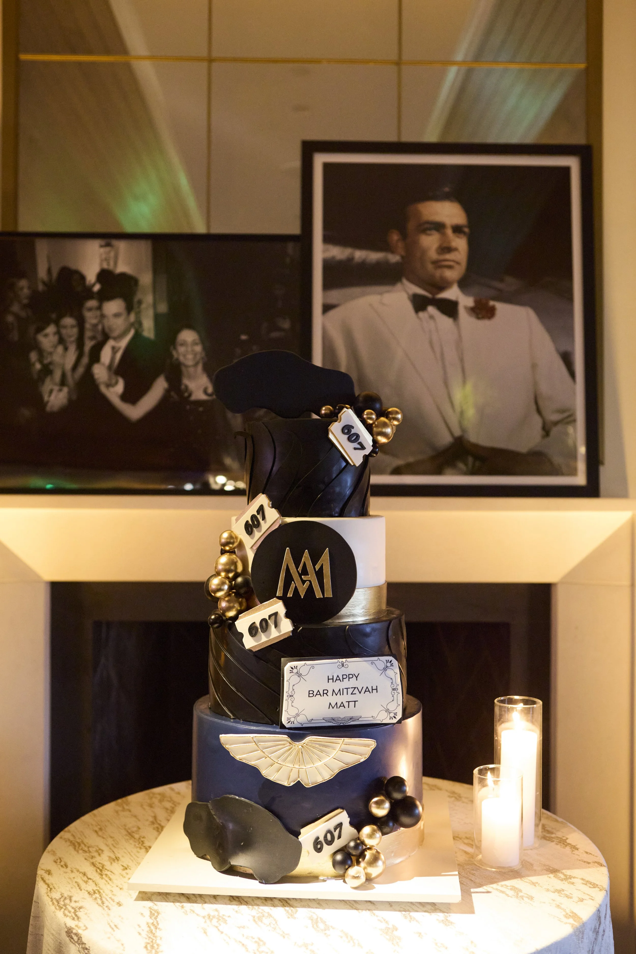 Black, white, and gold three-tiered cake with M1 logo, gold balls, and a white ornament, celebrating a Bar Mitzvah for Matt, with photos of a man in a white tuxedo in the background and candles on the table.