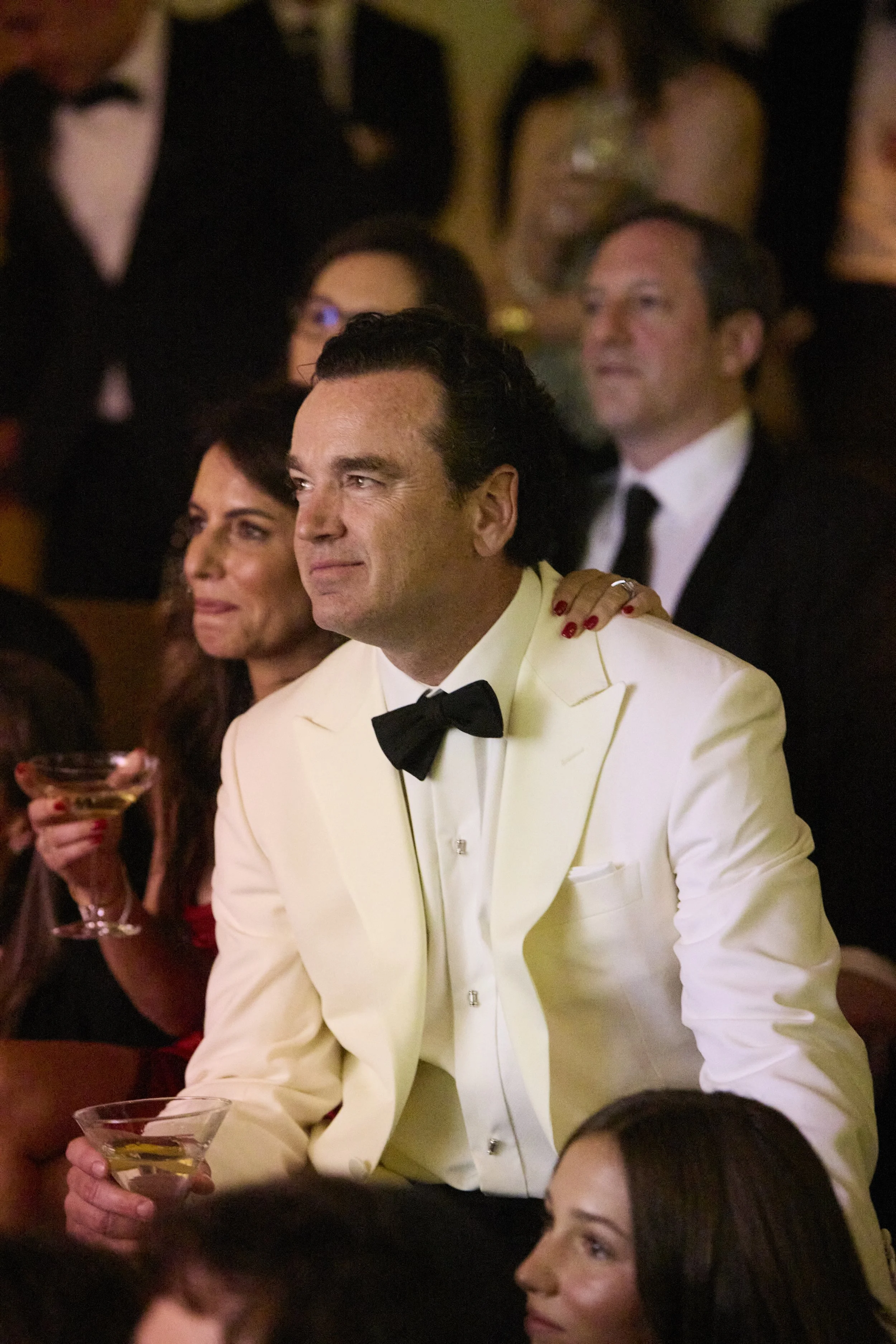 A man in a white tuxedo with a black bow tie holding a martini glass, sitting among a crowd at a formal event.