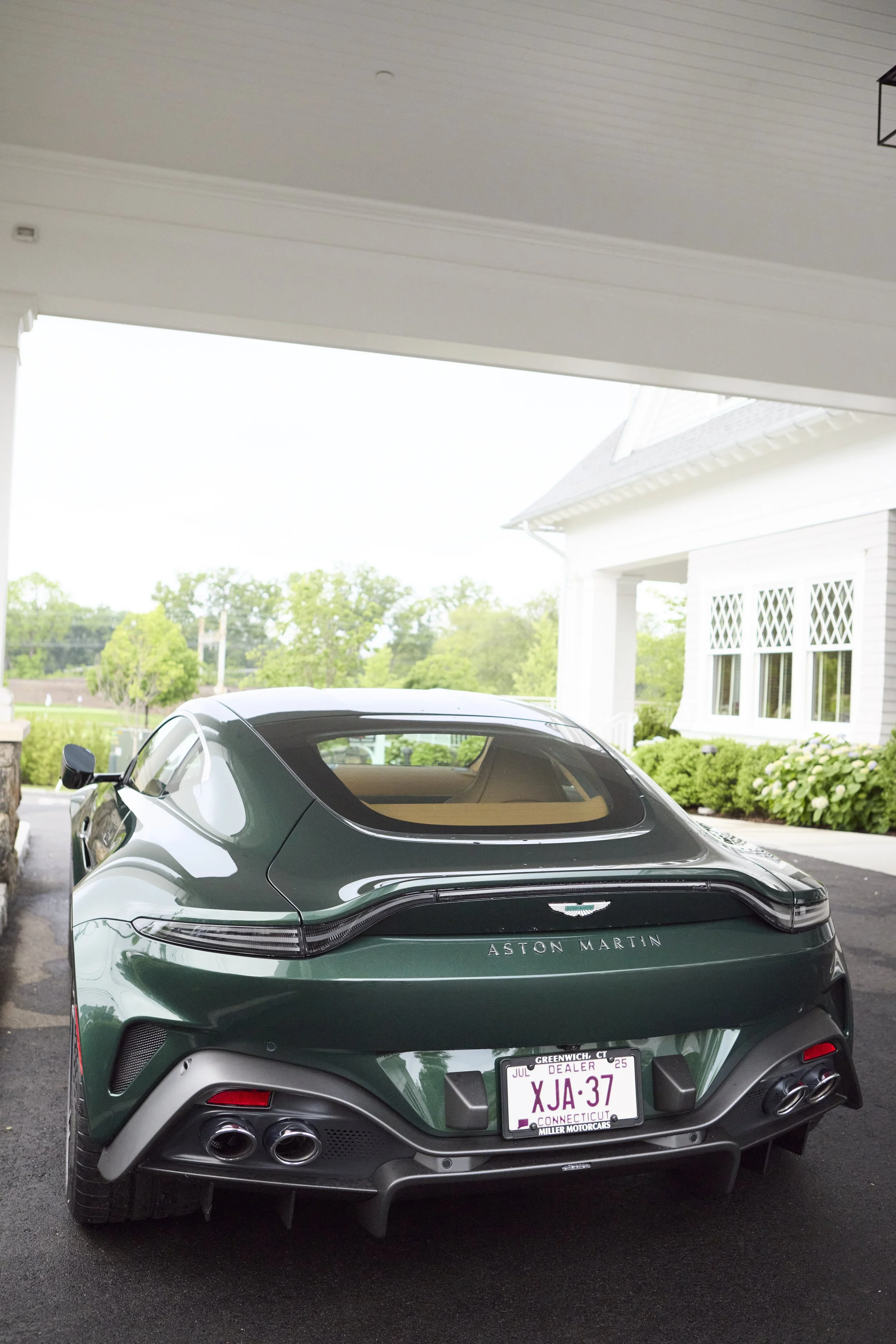 Green Aston Martin sports car parked in a residential driveway with white house and green trees in the background.