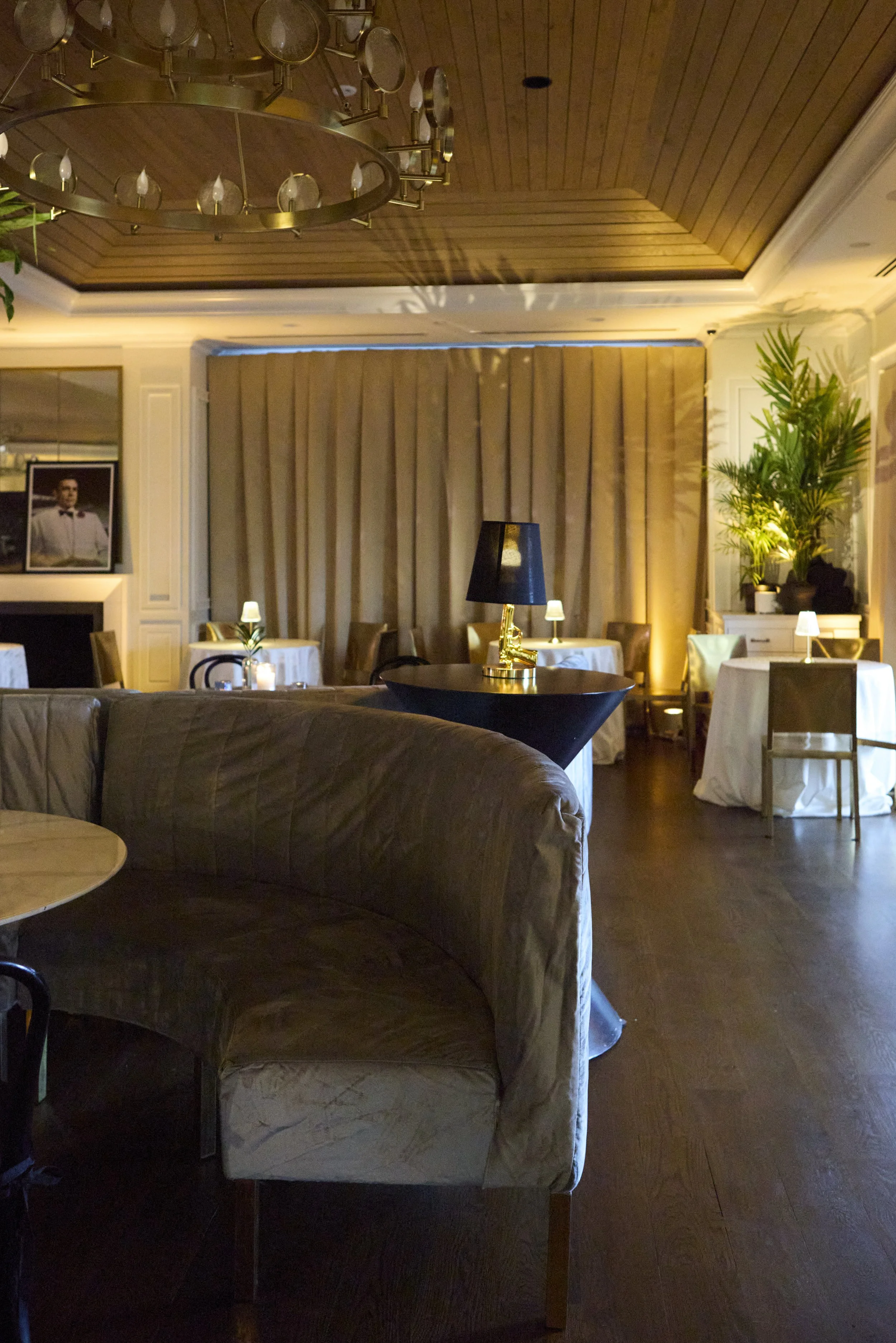 Elegant restaurant interior with wood-paneled ceiling, chandelier, and dim lighting. Features round tables with white tablecloths, small lamps, and tall green plants.