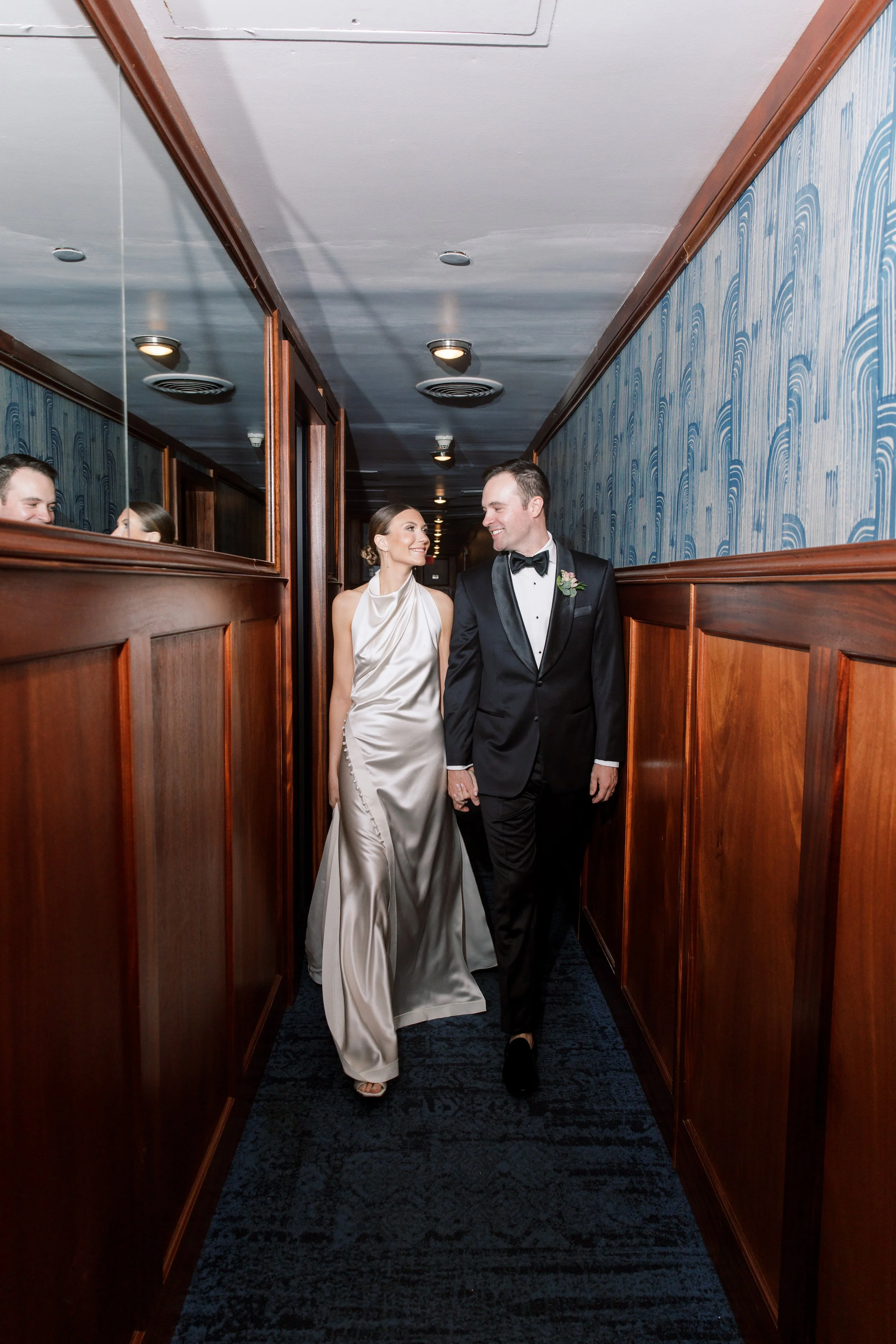 A bride and groom walking hand in hand down a corridor, smiling at each other. The bride is wearing a satin wedding gown, and the groom is dressed in a tuxedo with a bow tie.