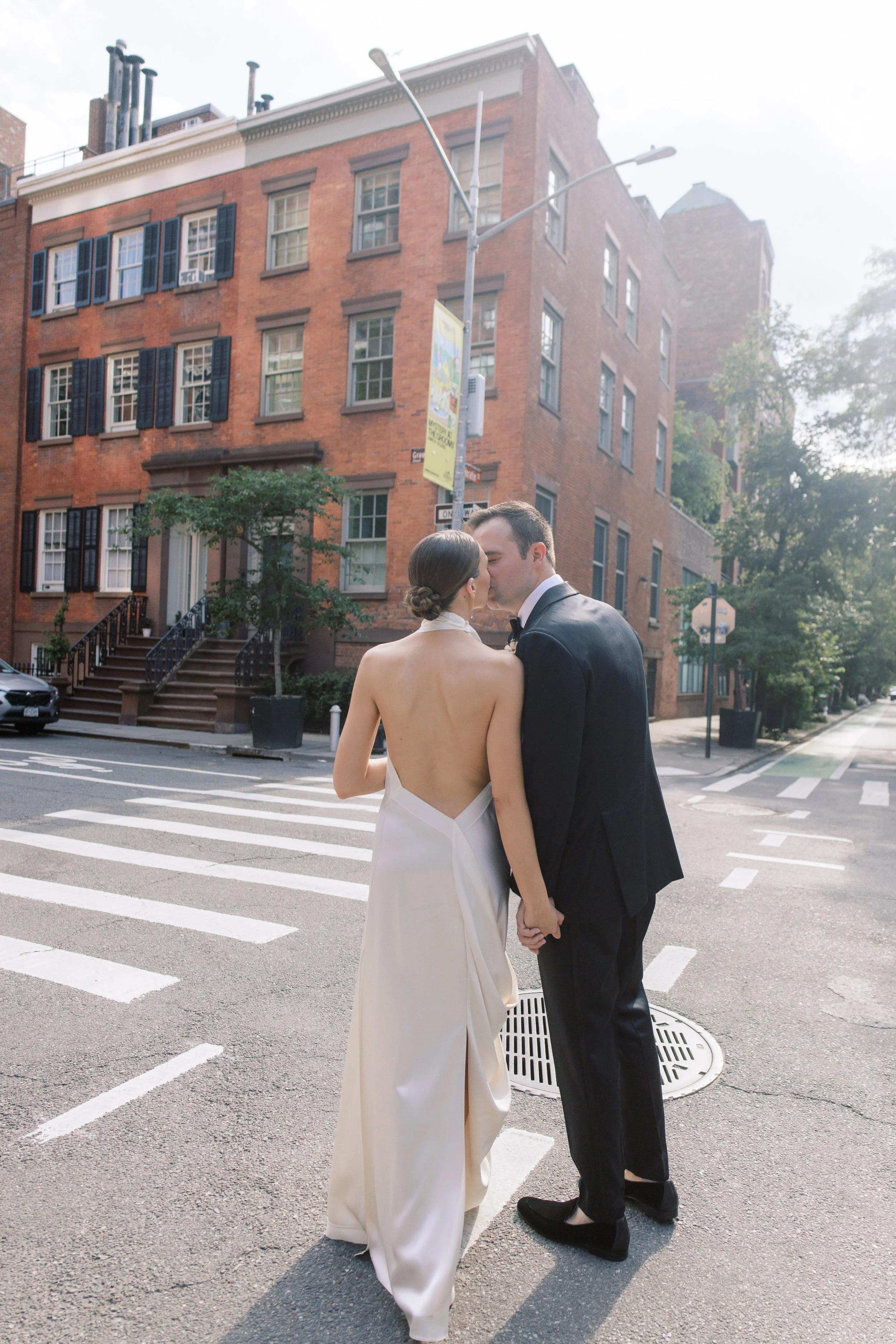 Elegant wedding couple standing along a tree-lined street in Chelsea NYC outside the Altman Building with red umbrellas and city skyline.
