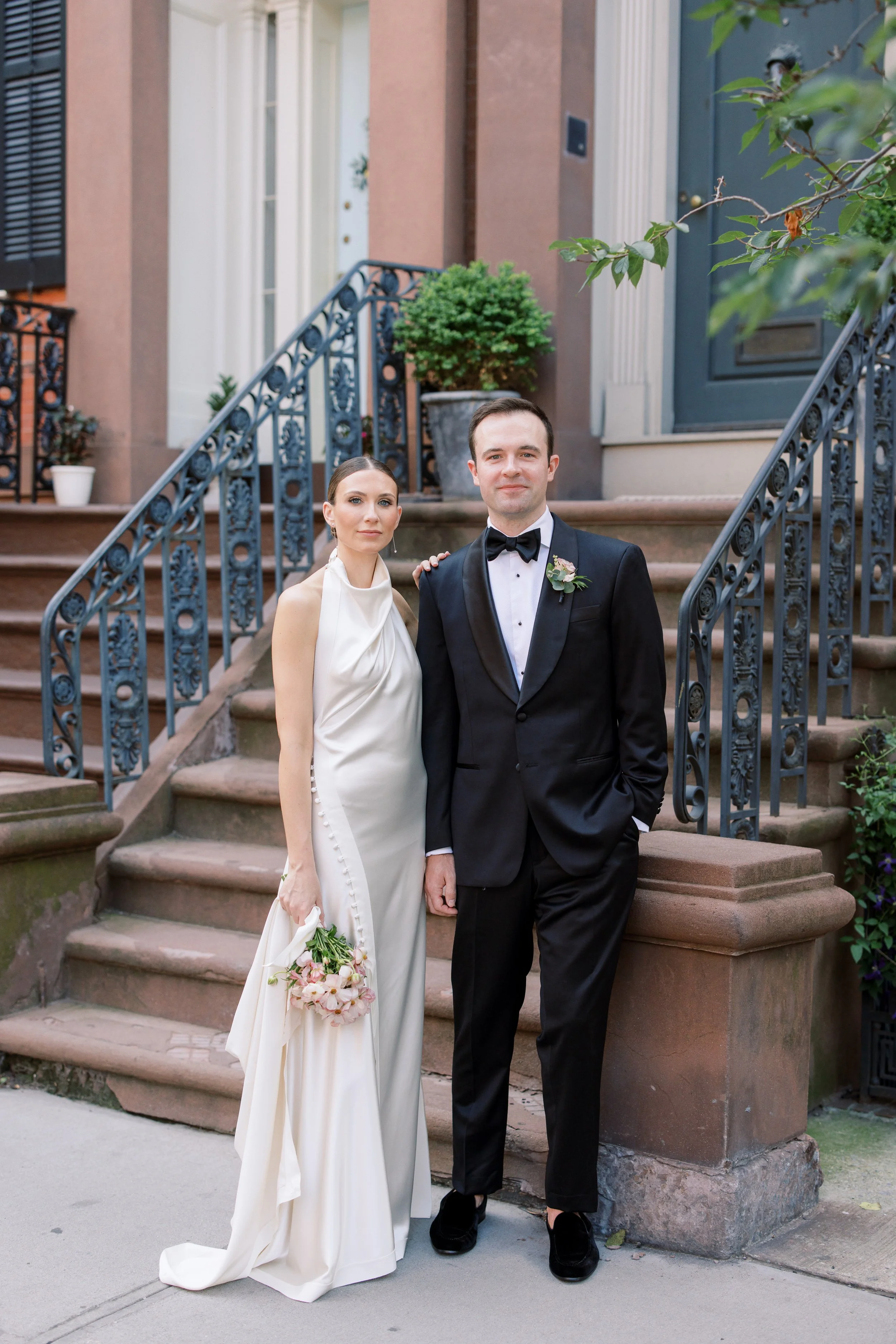 Elegant wedding couple standing along a tree-lined street in Chelsea NYC outside the Altman Building with red umbrellas and city skyline.