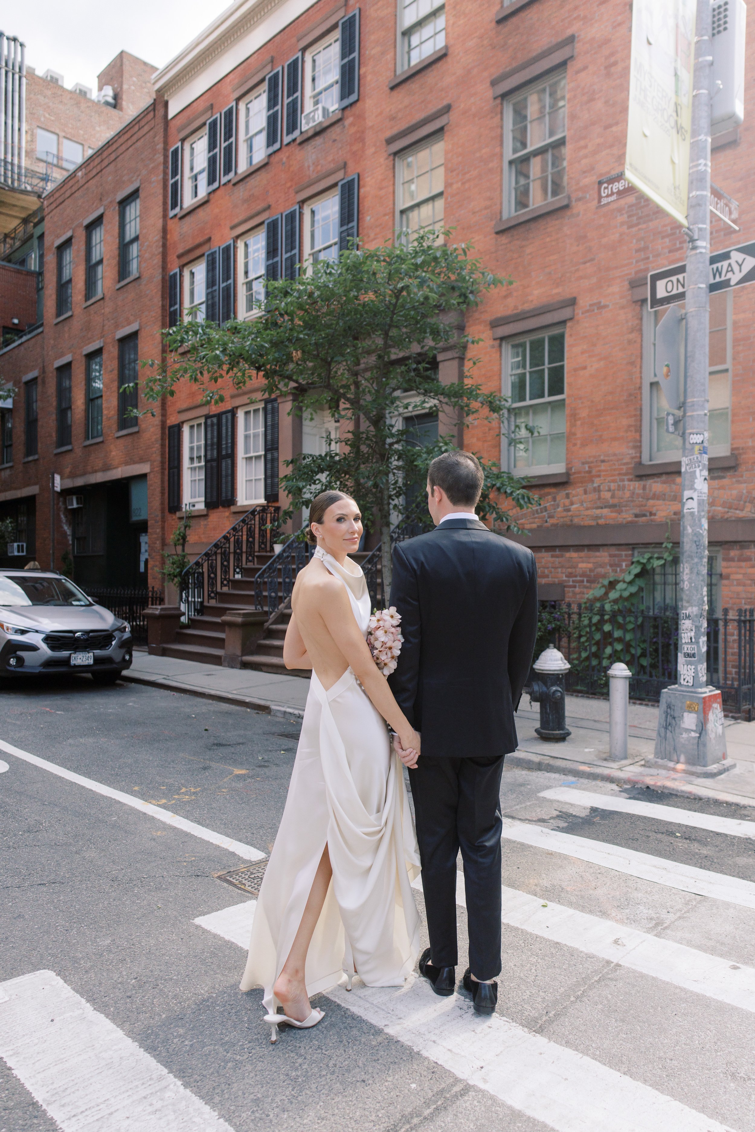 Elegant wedding couple standing along a tree-lined street in Chelsea NYC outside the Altman Building with red umbrellas and city skyline.