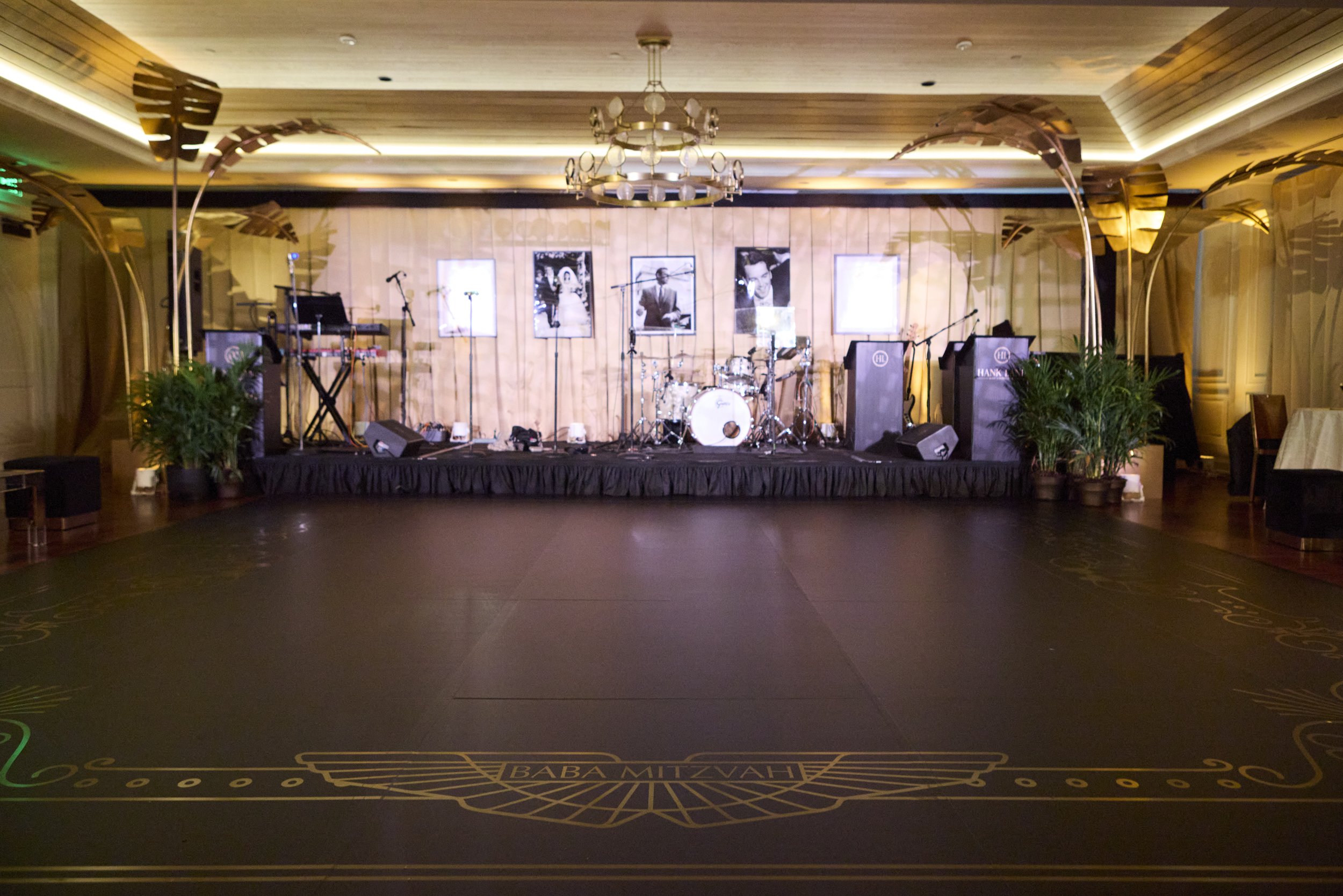 Empty stage set up with musical instruments, including drums, keyboard, and microphones, in a decorated ballroom with framed photos on the wall and potted plants on each side.