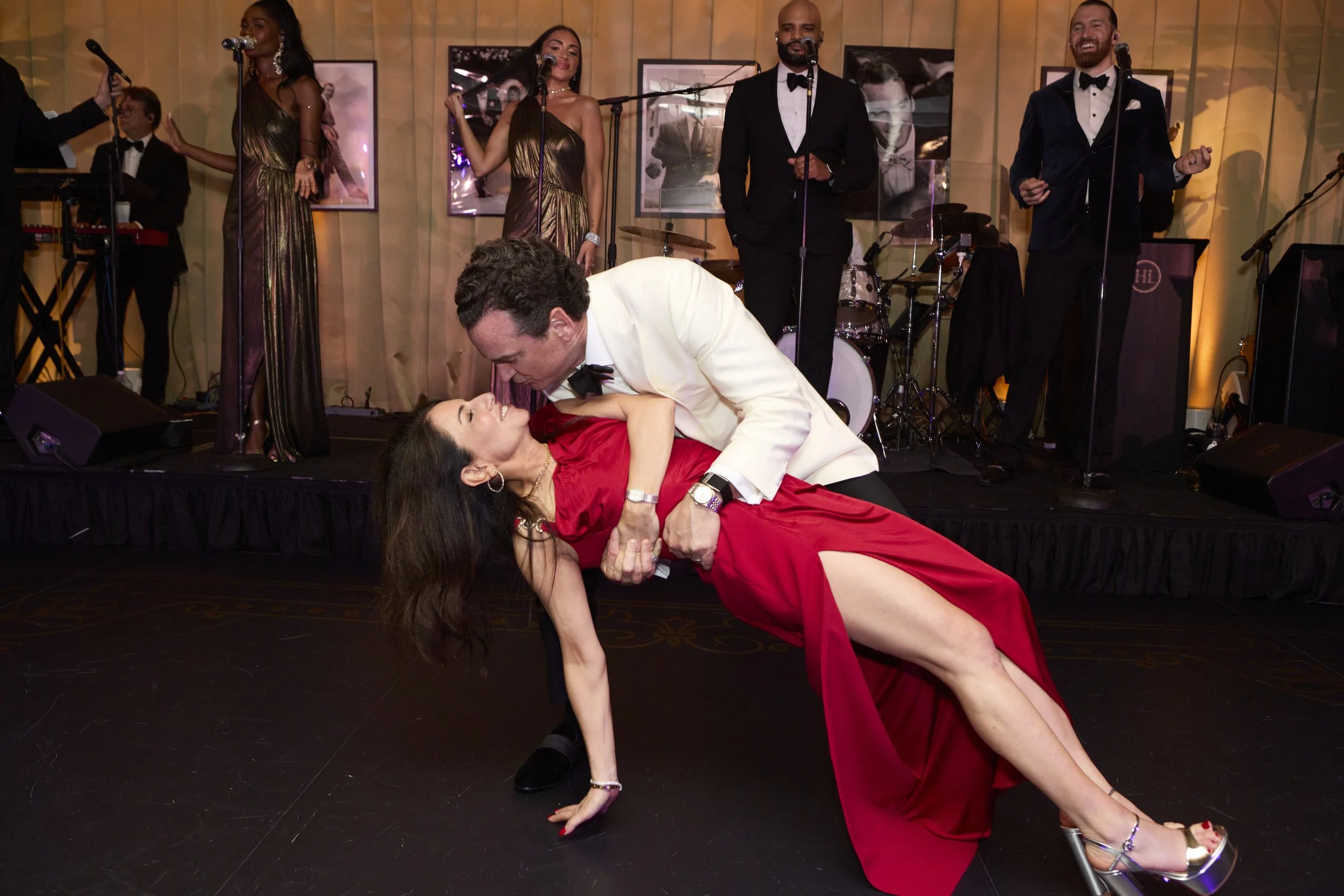 A man and woman dance closely at a formal event with a live band performing on stage in the background.
