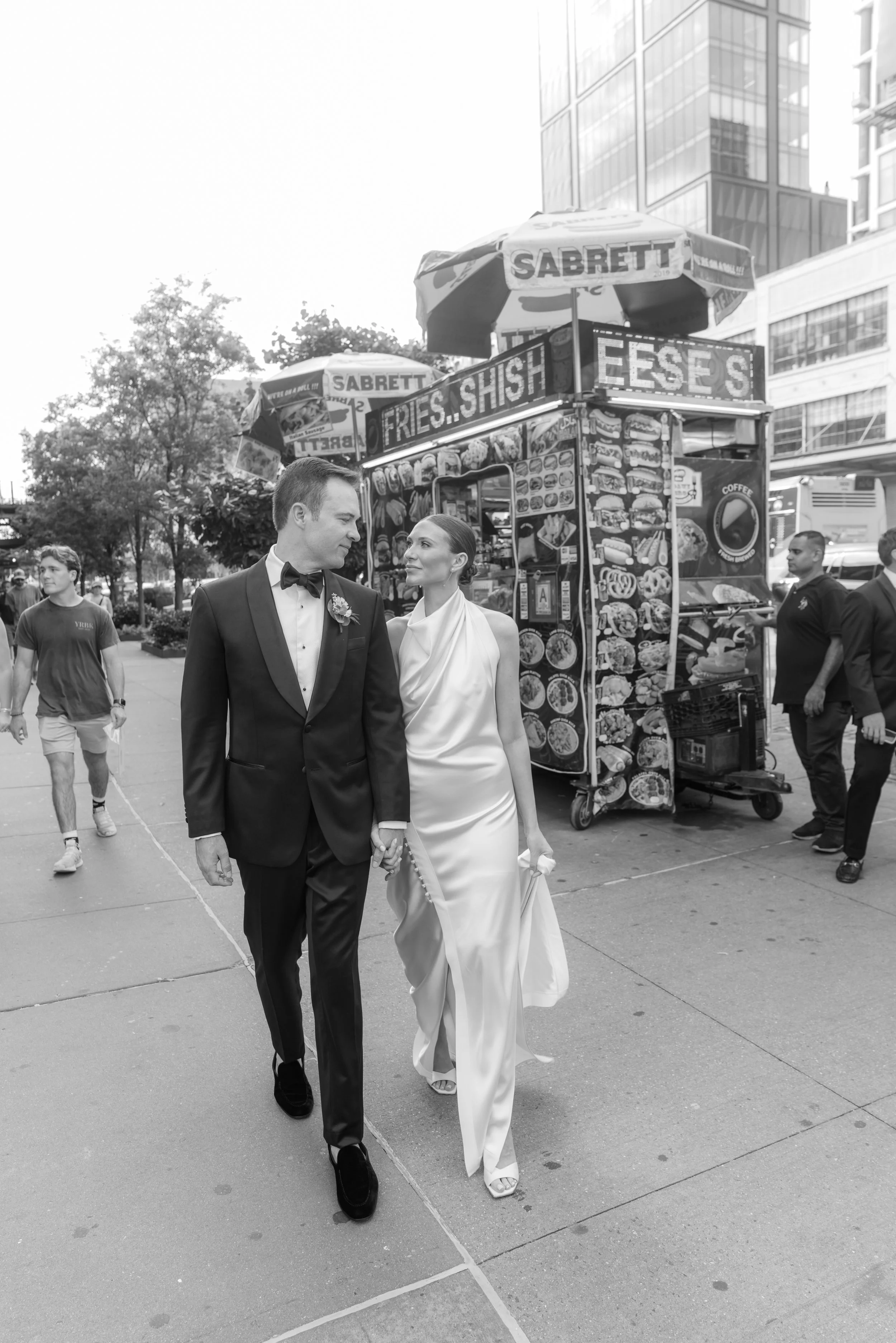 A couple dressed in formal wedding attire walking hand in hand on a city sidewalk, with a hot dog stand in the background.