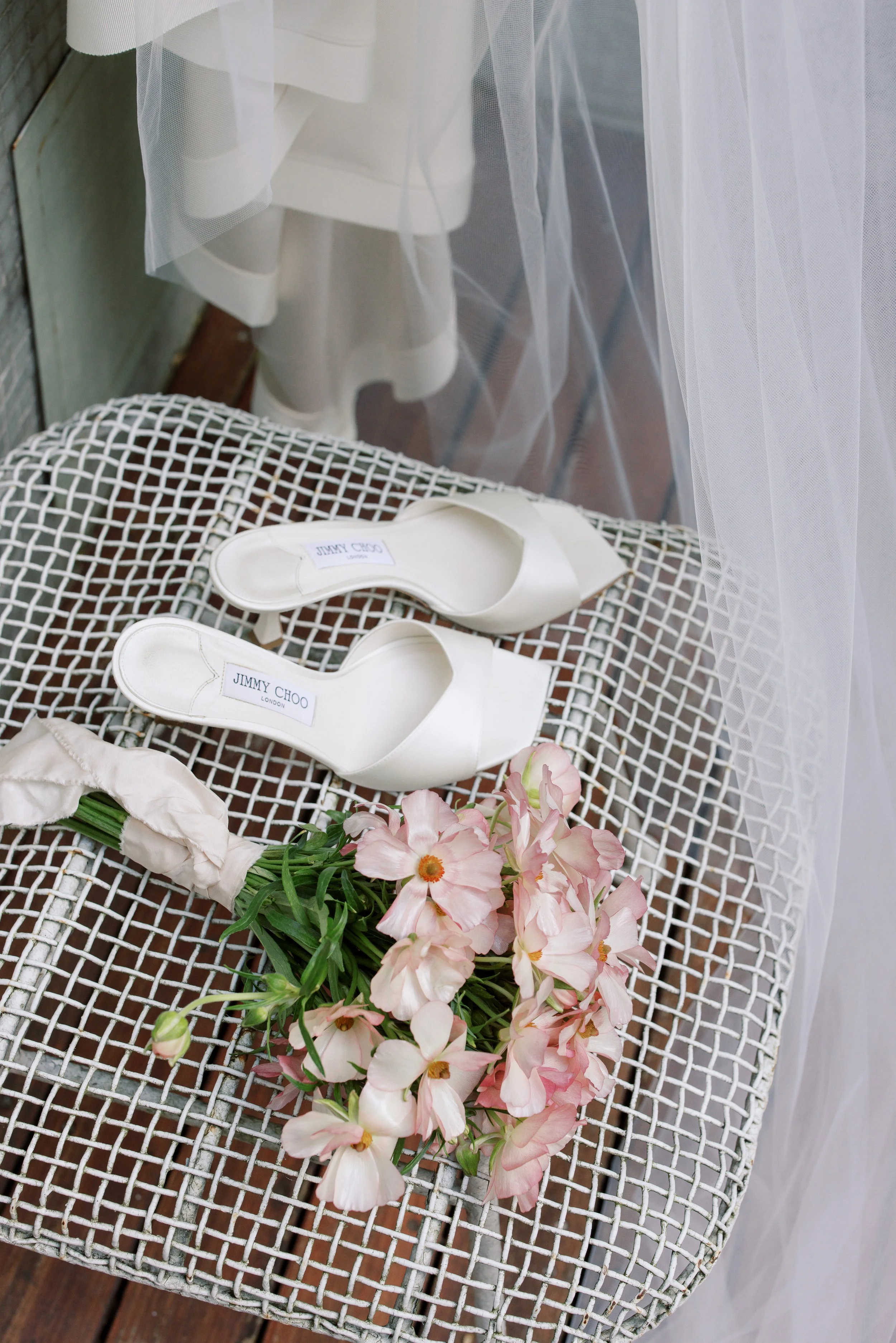 A pair of white Jimmy Choo heels placed on a white woven chair, with a bouquet of pink and white flowers next to them, near a window with sheer white curtains.
