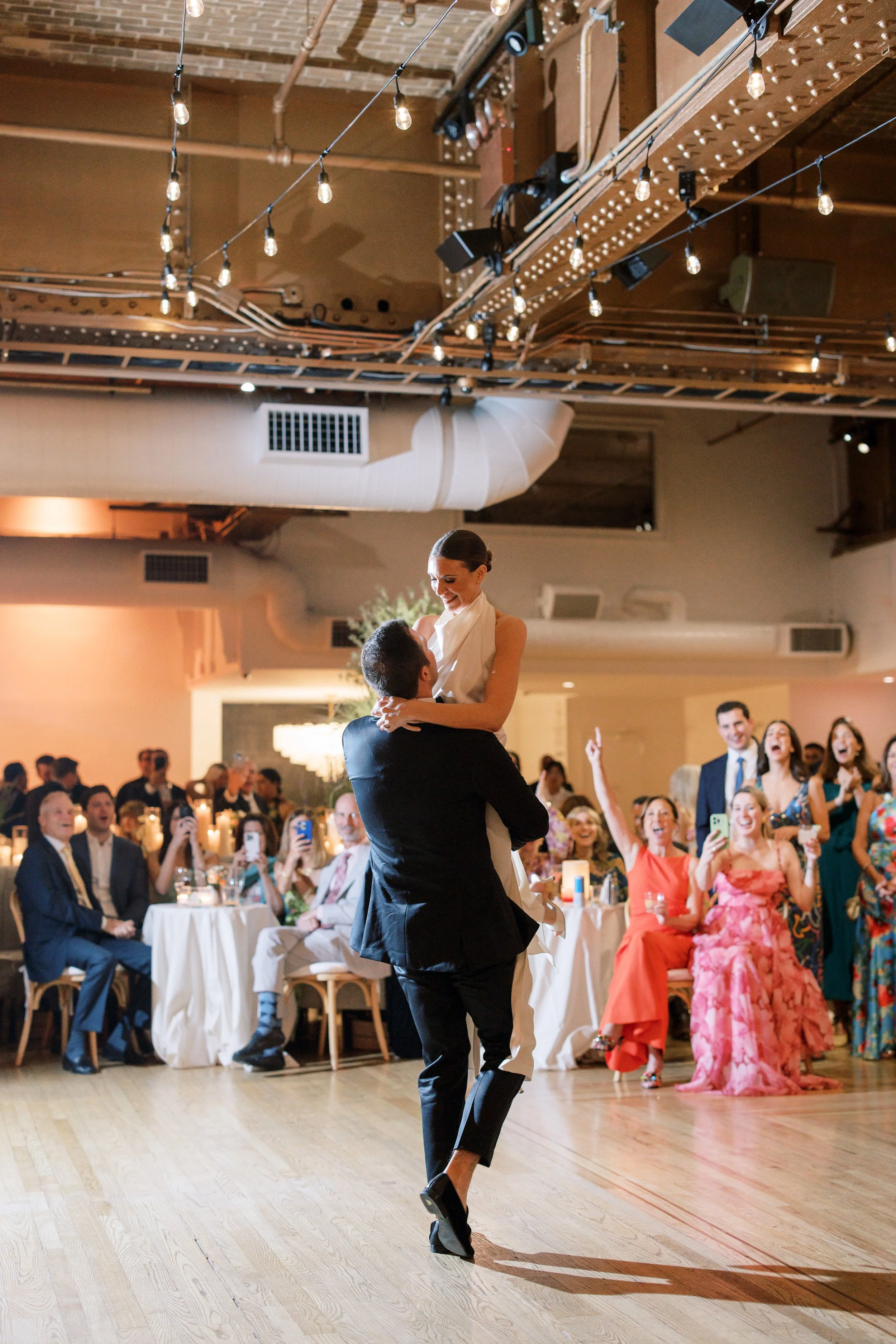 A bride and groom dance at their wedding reception. The groom lifts the bride while she smiles, surrounded by seated and standing guests who are taking photos and videos, clapping, and celebrating.
