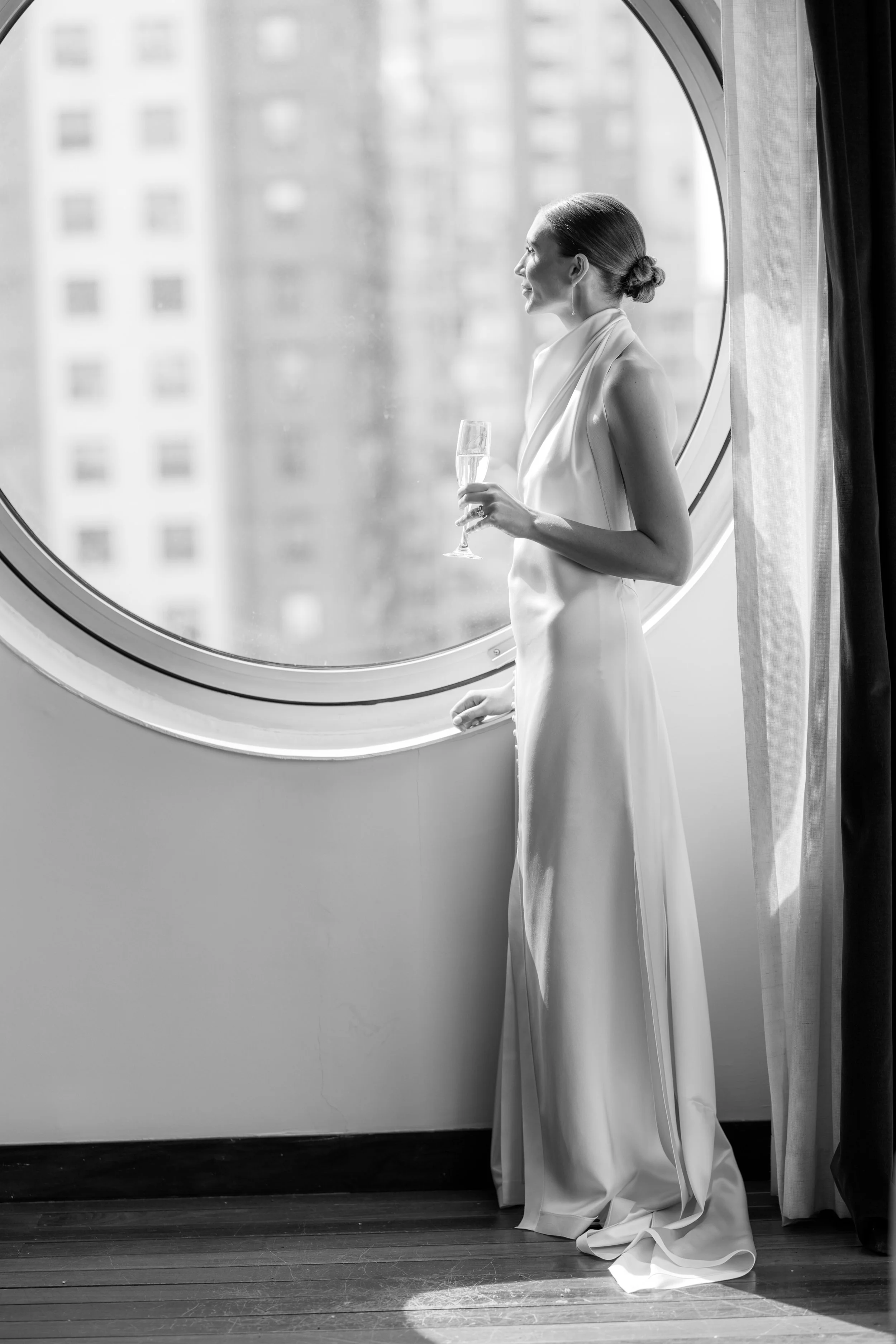 A woman in a long white dress standing by a large circular window, holding a champagne flute, looking outside.