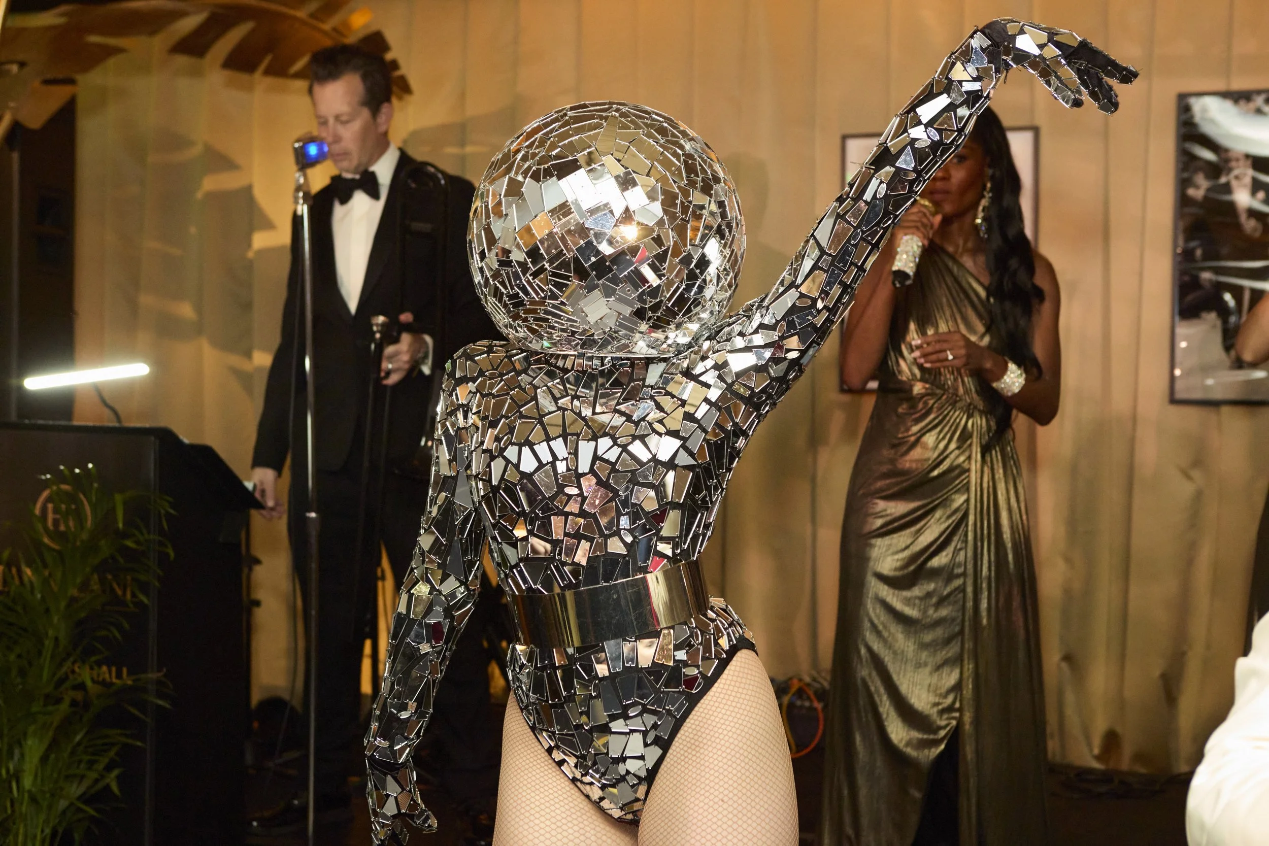A person in a mirrored mosaic costume with a disco ball head, posing with one arm raised. Two people are in the background, one in a tuxedo and the other in a gold dress, at an indoor event.