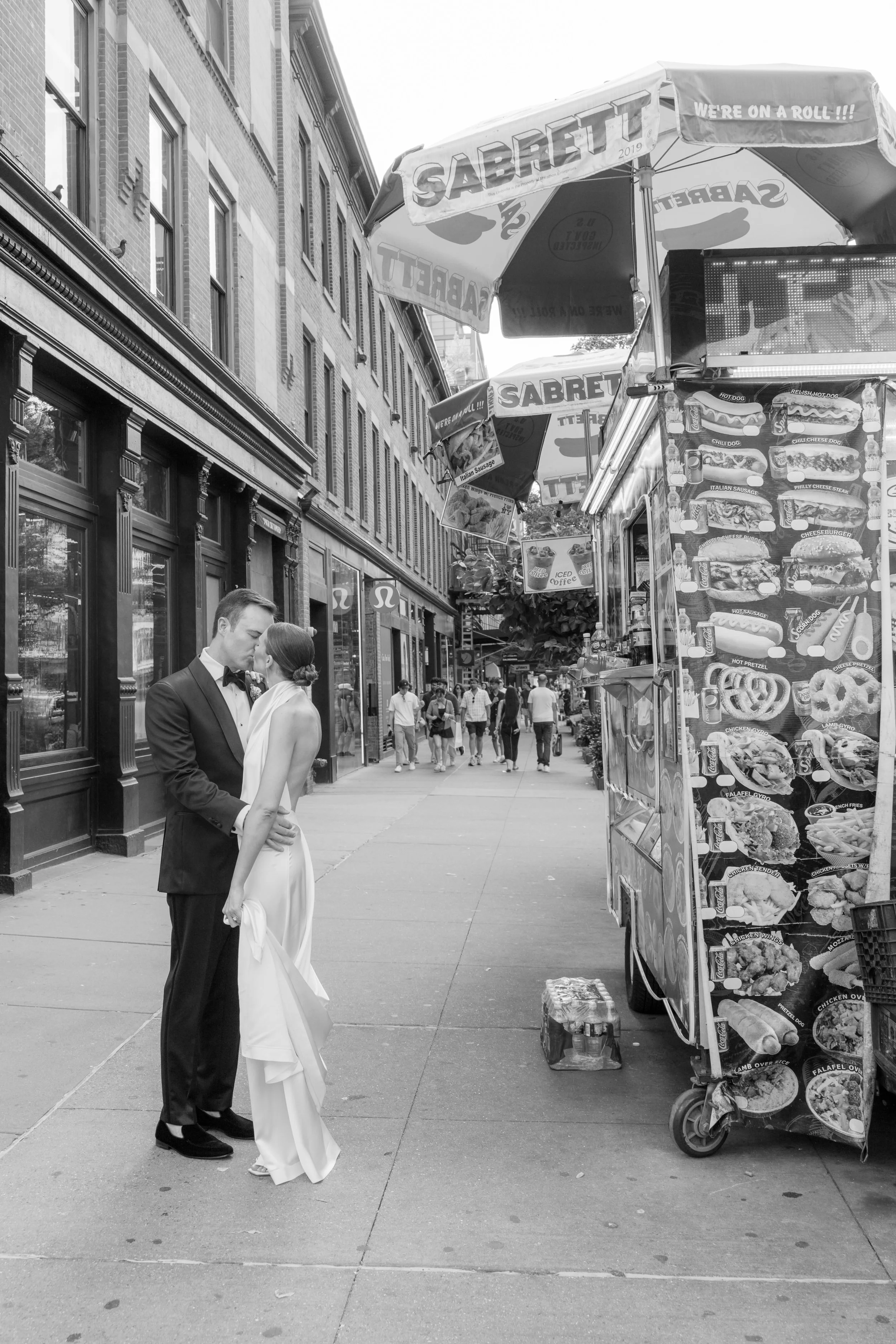 A couple in wedding attire sharing a kiss on a city sidewalk, with a food stand on the right and people walking in the background.