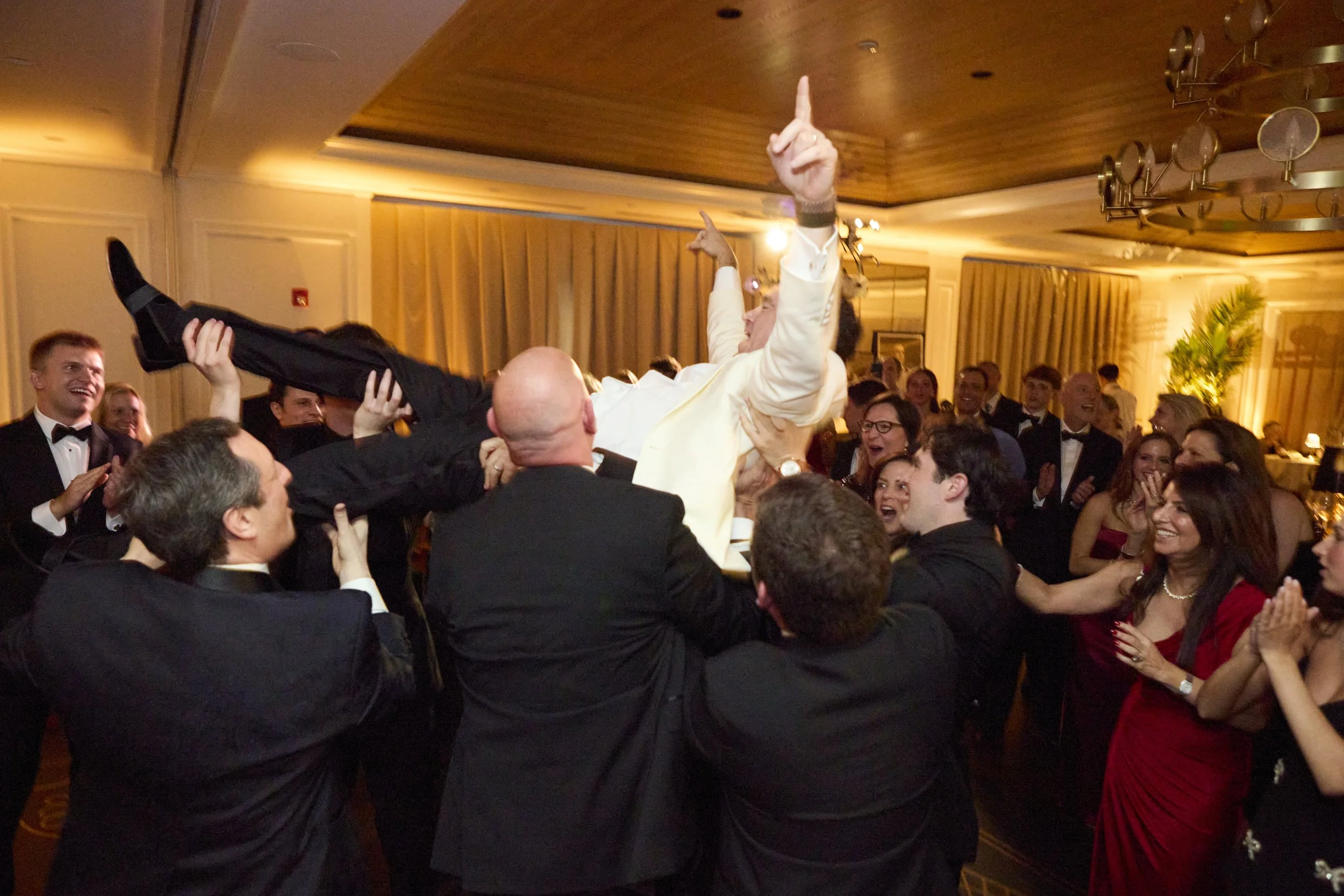 A celebration at a formal event with a group of people lifting an individual in a white suit into the air, surrounded by other attendees in tuxedos and evening gowns, in a decorated room with warm lighting.