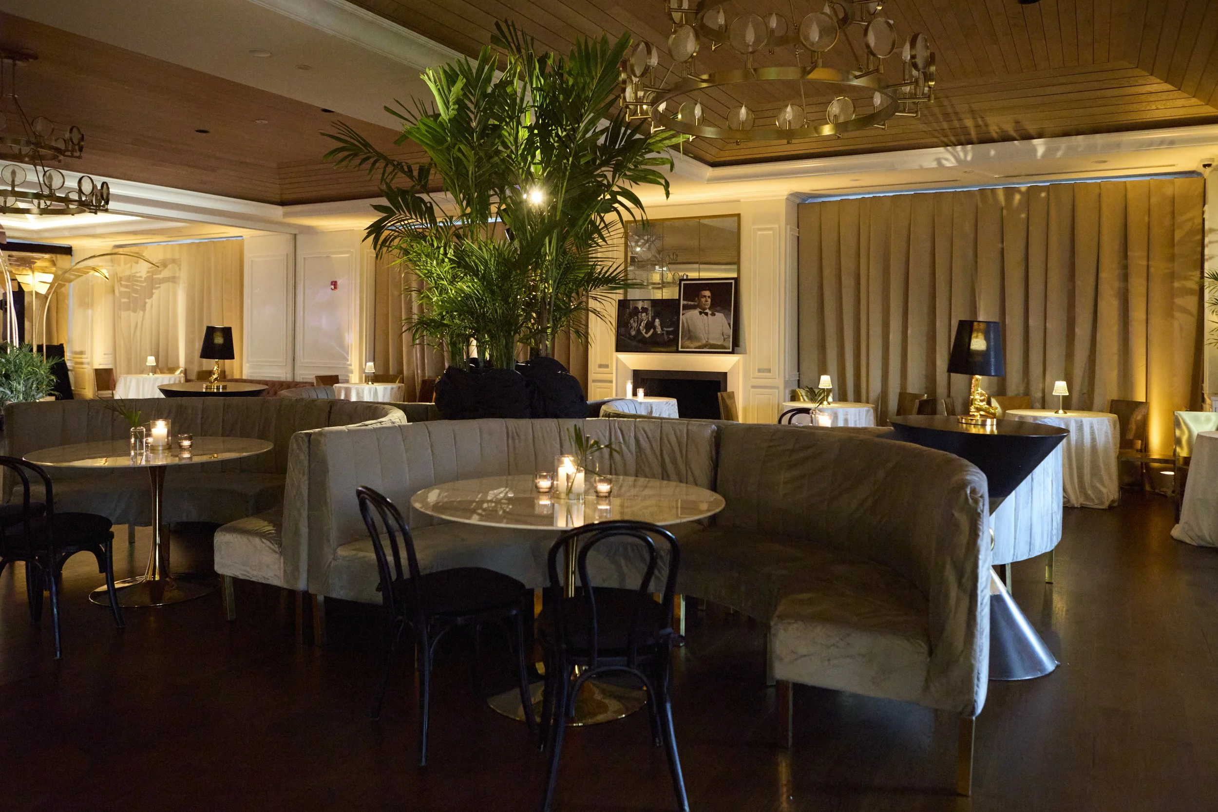 Elegant restaurant interior with round tables, candles, and black and white lampshades; large green plant in the center; framed black and white photos above a fireplace; warm lighting, dark wooden floors, beige curtains, and a wood-paneled ceiling.