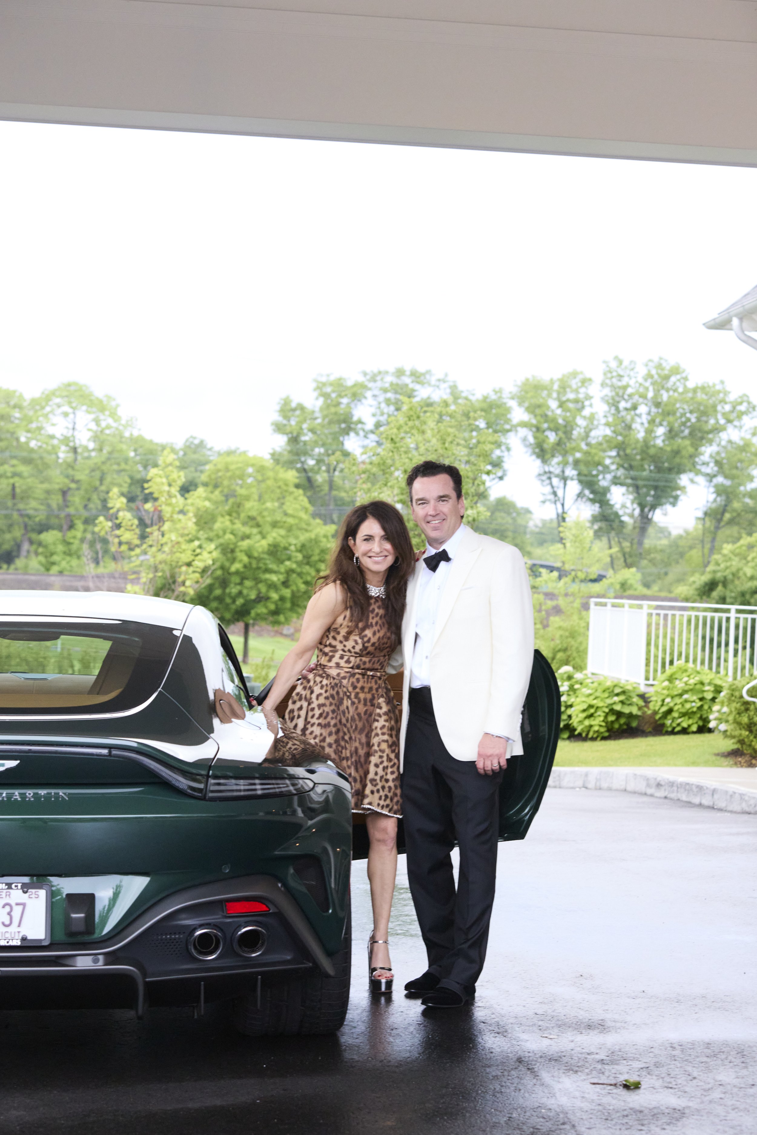 A man and woman dressed in formal attire standing outside next to a black luxury car with a green landscape in the background.
