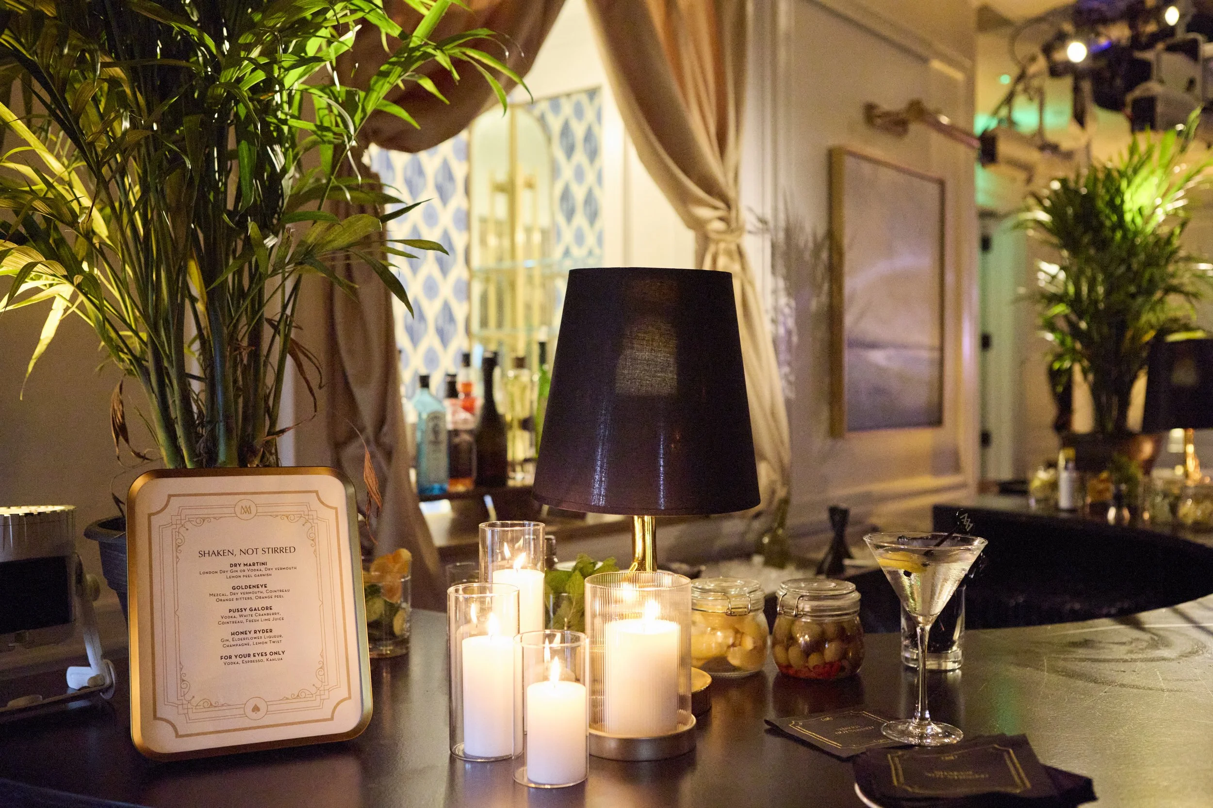A cozy bar or lounge area with table lit by candles, a black-lamp with a gold stand, jars of candies, a martini glass with an olive, and a menu with the title 'Shaken, Not Stirred'.