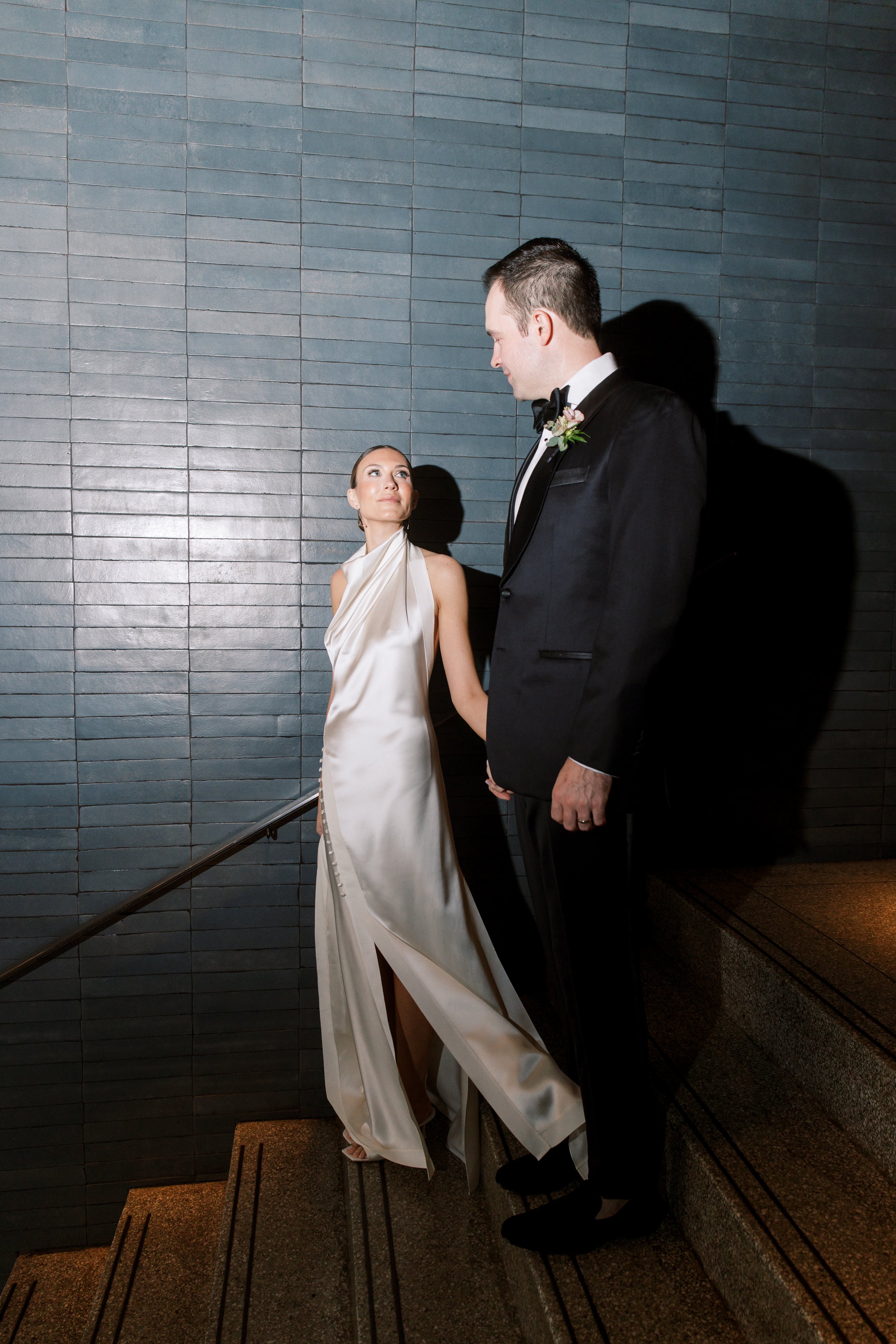 Bride and groom standing on staircase, holding hands, looking at each other, against a dark tiled wall, bride in a satin gown, groom in a tuxedo with bow tie and boutonniere.