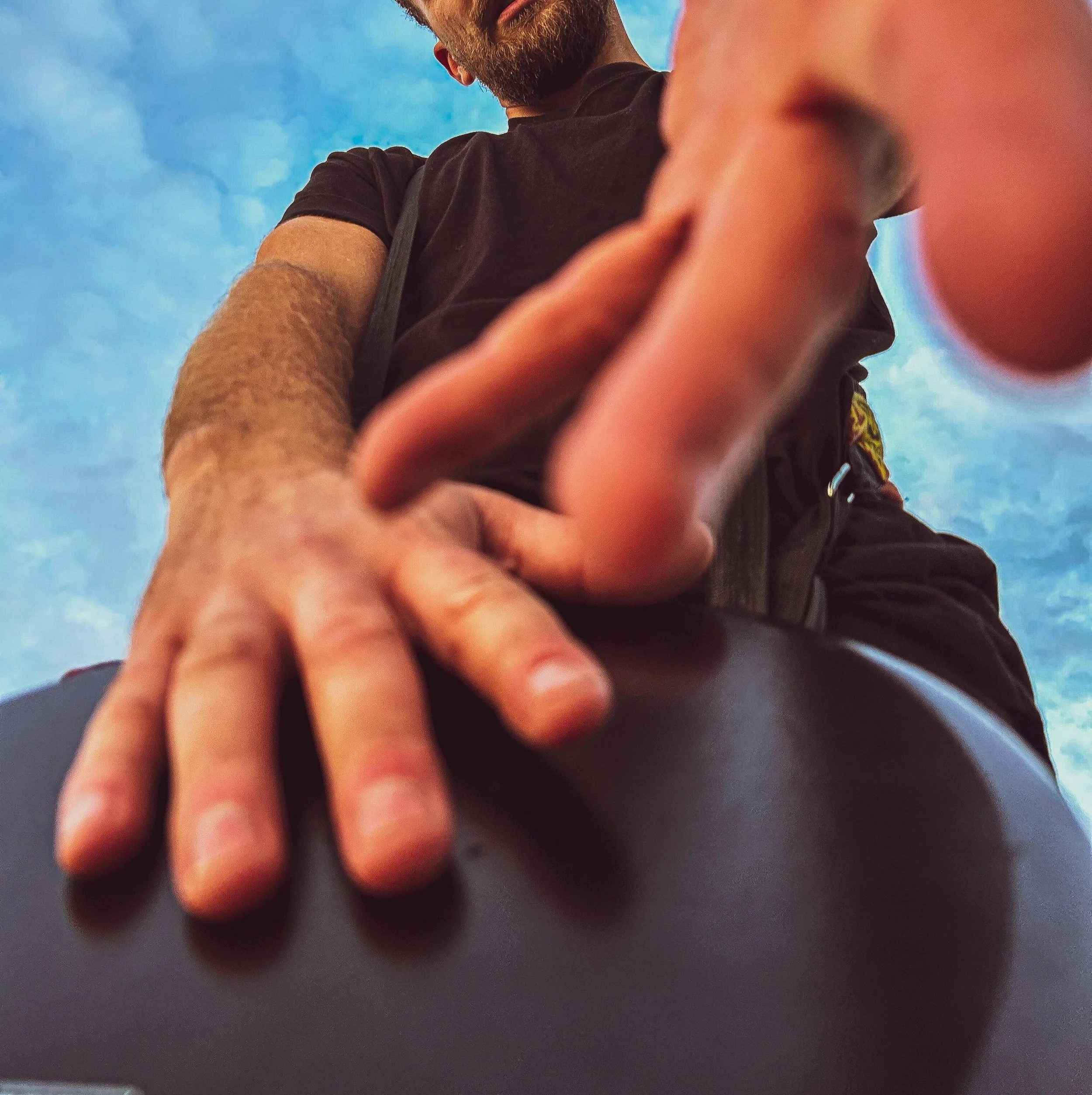A person reaching towards the camera with their hand, set against a blue sky with some clouds. Tobi Ripley. Roaming bongo drummer. Percussionist. event drummer. wedding entertainment. festival drummer. dj sax percussion. drum player