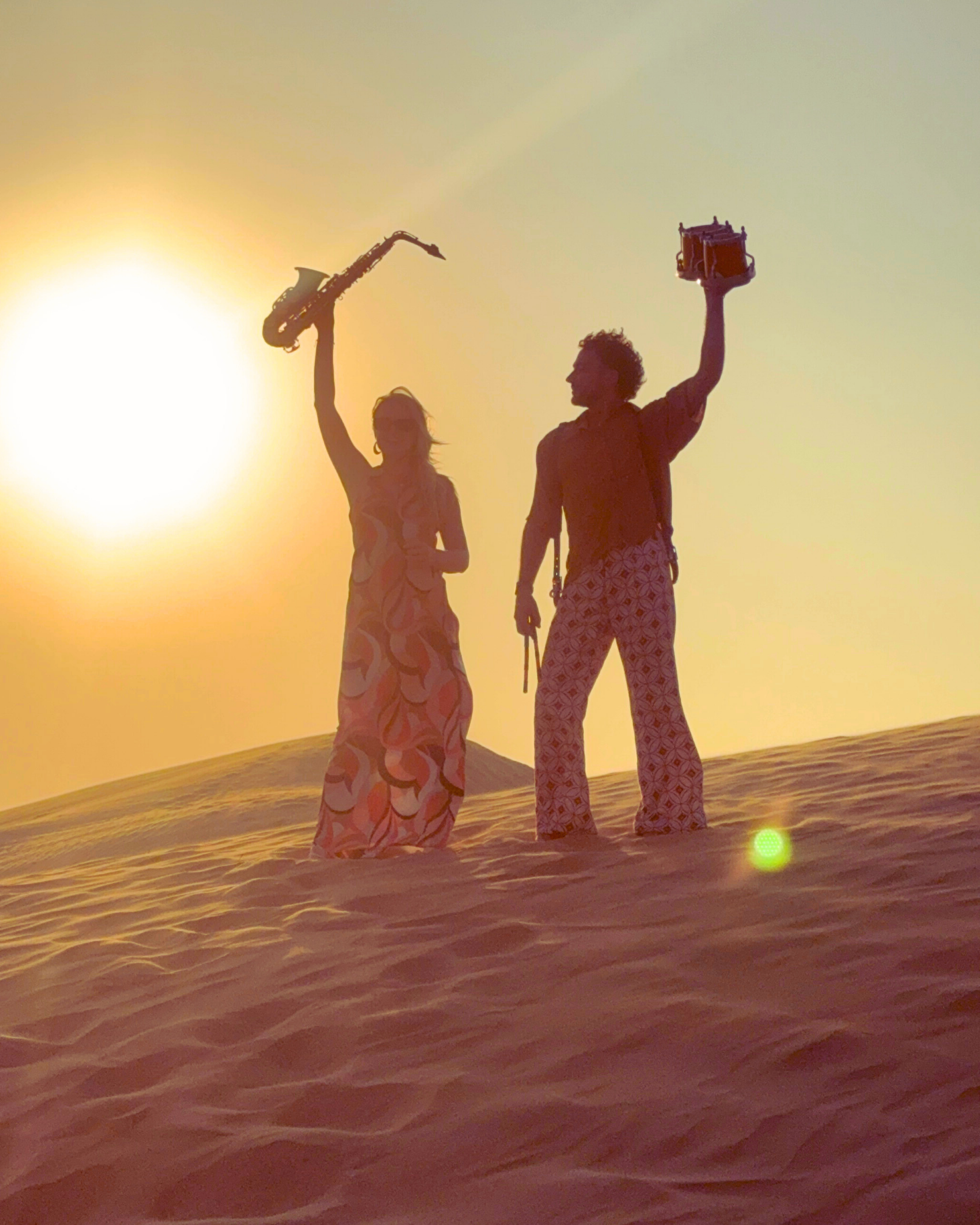 Two people silhouette standing in the desert at sunset, holding musical instruments and equipment, with the sun low in the sky.