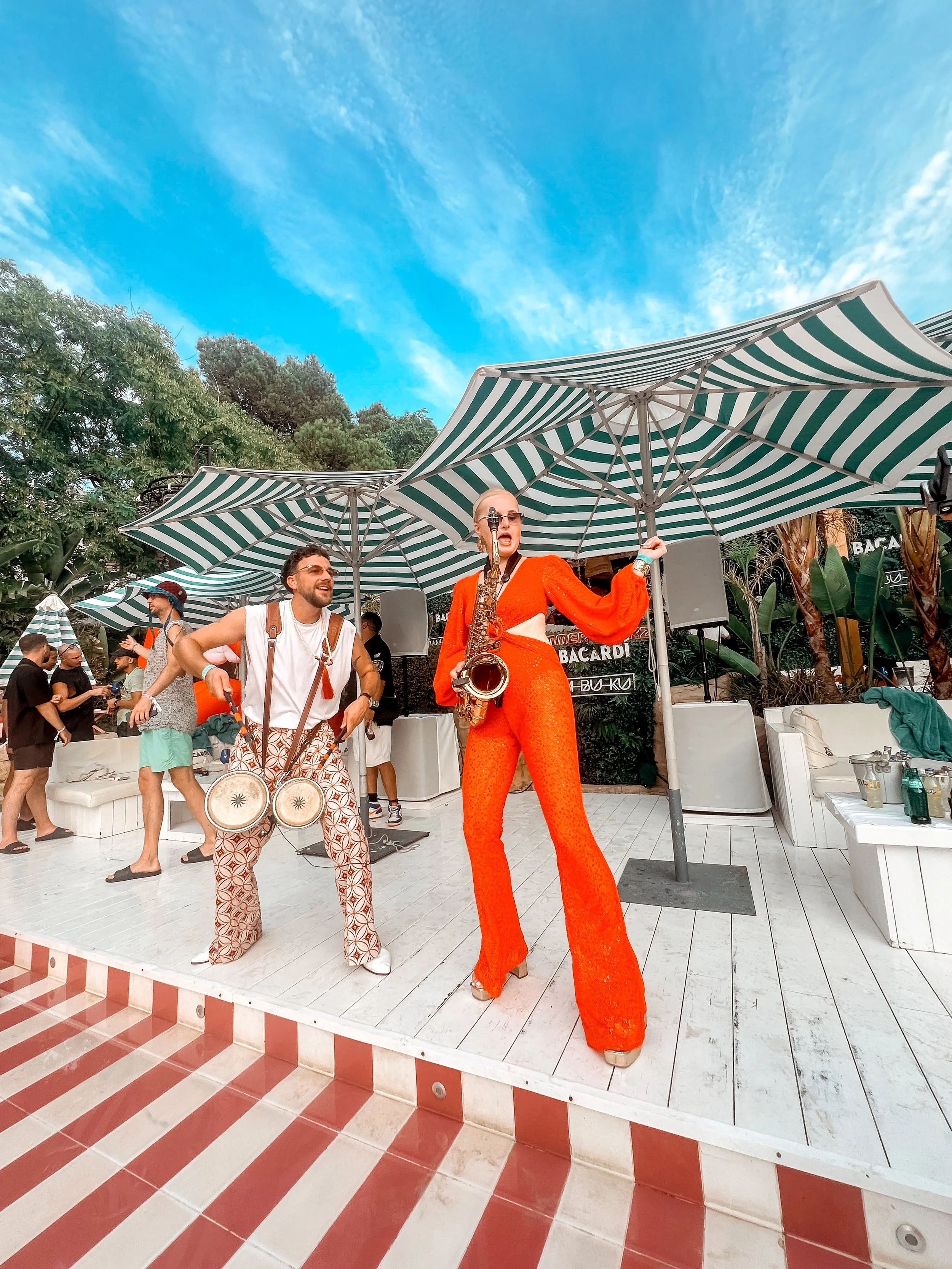 Two performers, a man and a woman, performing at an outdoor event. The woman is wearing an orange jumpsuit and playing a saxophone, while the man is dressed in patterned pants, a white shirt, and playing drums. They are under striped umbrellas with a lively crowd around them.