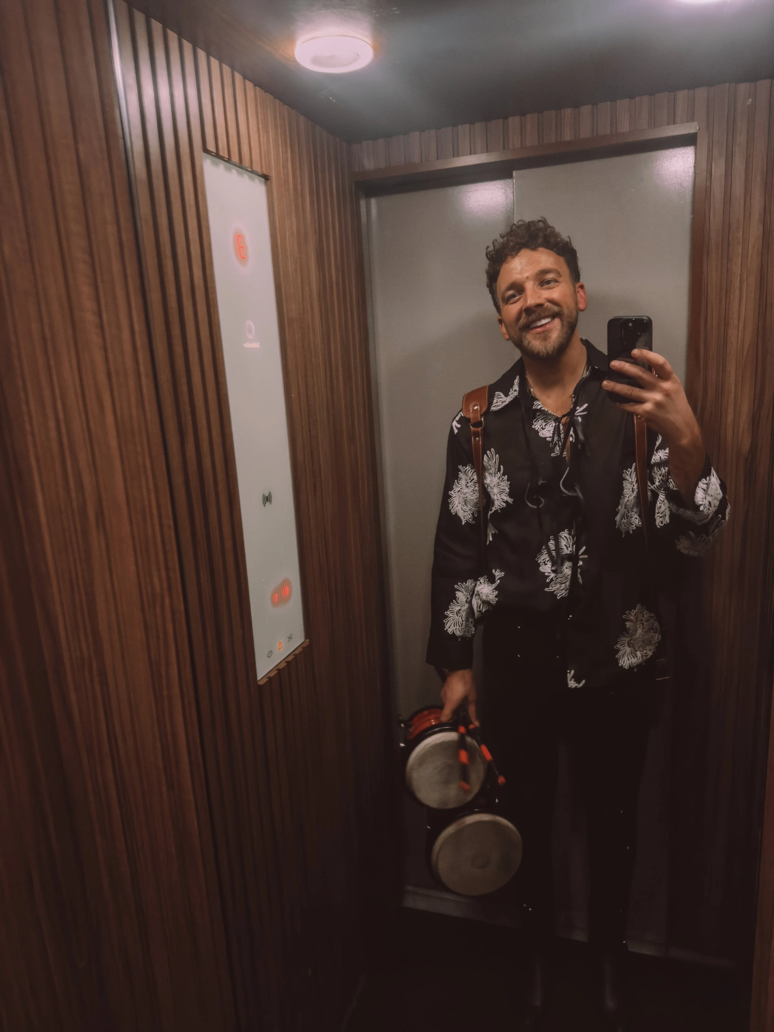A man with a beard and curly hair, smiling, taking a mirror selfie in an elevator with wooden wall panels. He is wearing a black shirt with white floral patterns, carrying a brown backpack, and holding a black and orange duffle bag. Tobi Ripley. Roam