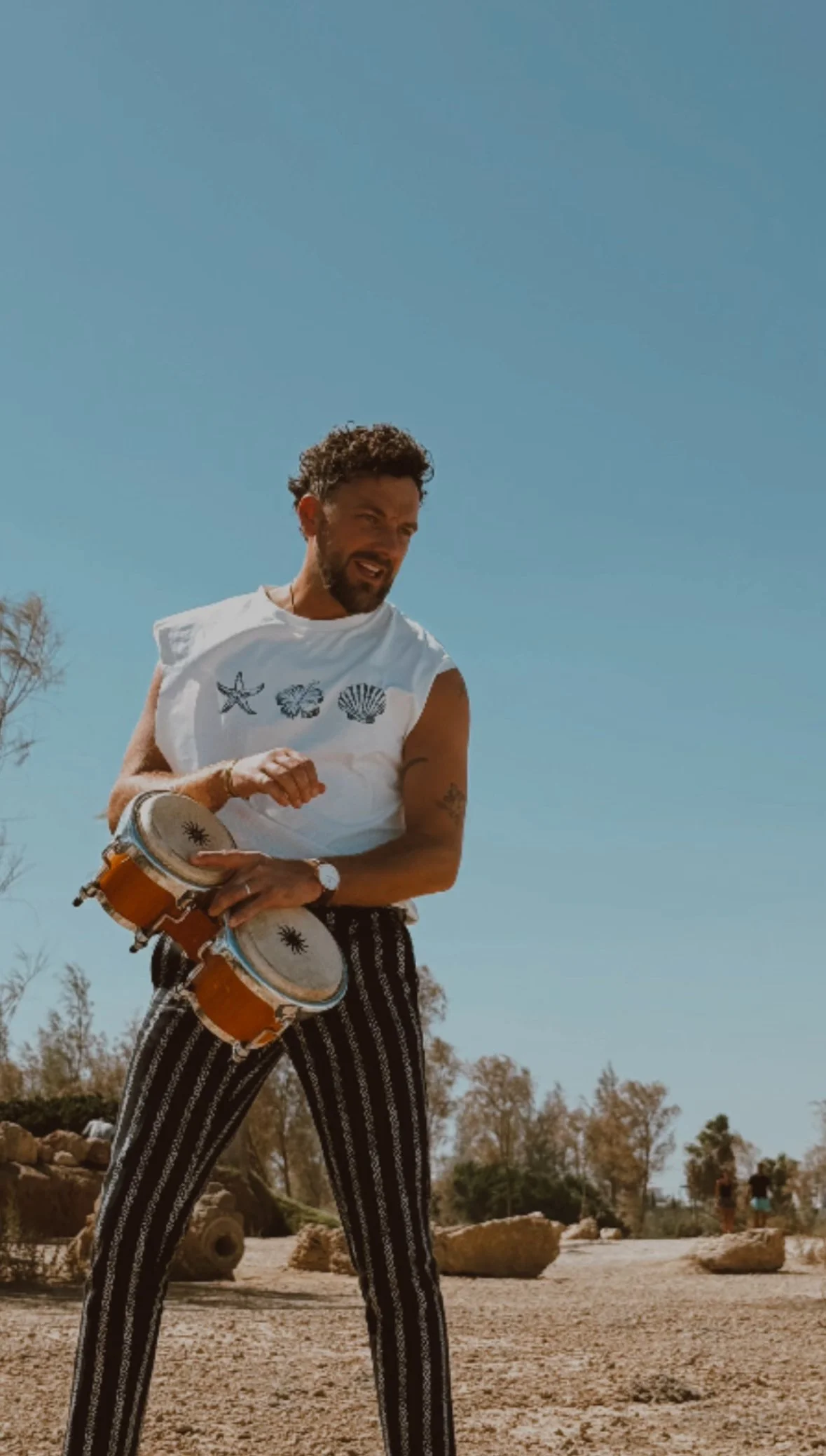 A man playing bongos outdoors during daytime, wearing a white sleeveless shirt with sea shell illustrations and striped black and white pants.