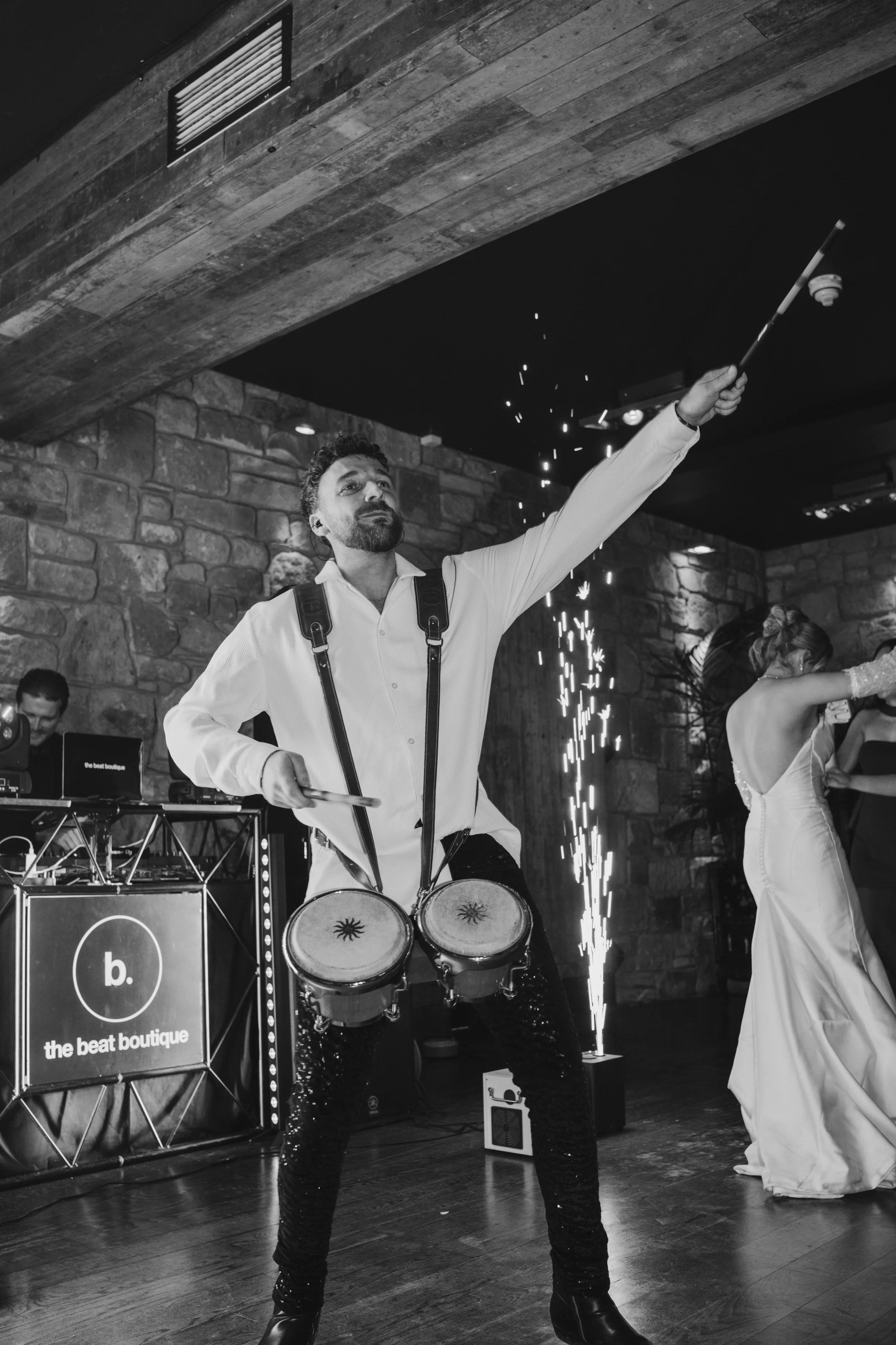 A man in a white shirt playing drums with percussion instruments hanging from his neck at a celebration, with a DJ in the background and a woman in a white dress dancing near a sparkler on the floor.