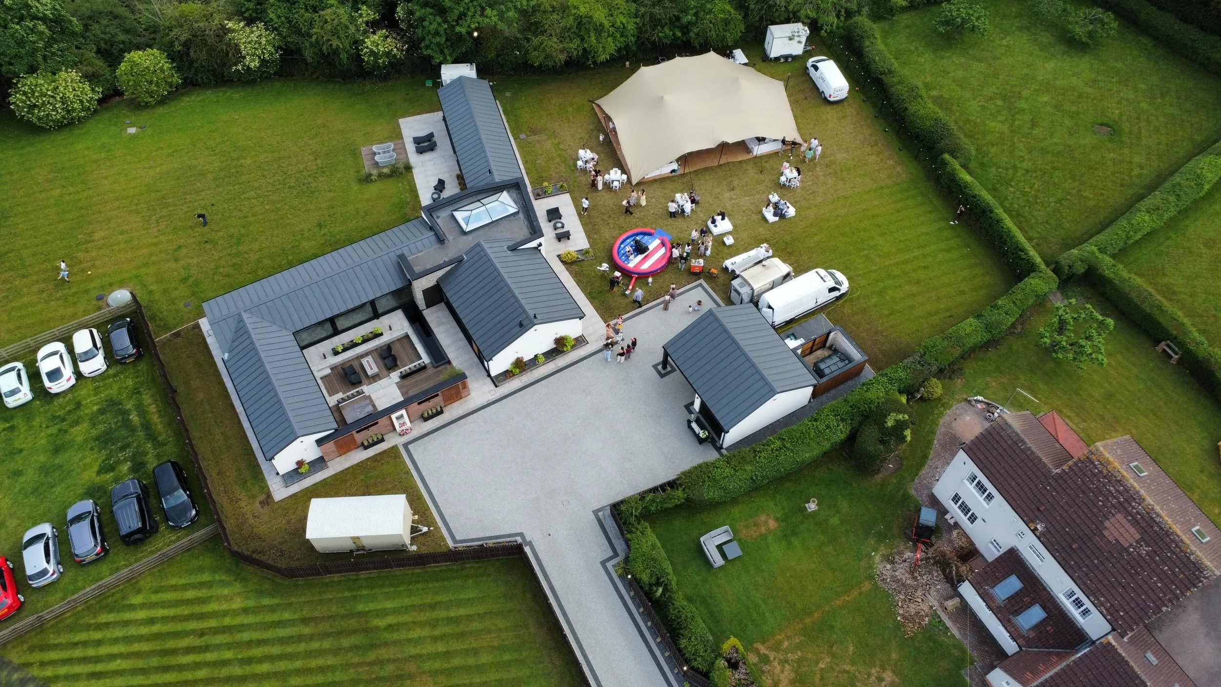 Aerial view of a backyard gathering with a beige canopy tent, people sitting at tables, a small inflatable pool, and multiple vehicles parked nearby. featuring TOBI RIPLEY on bongos roaming percussionist international dj drummer